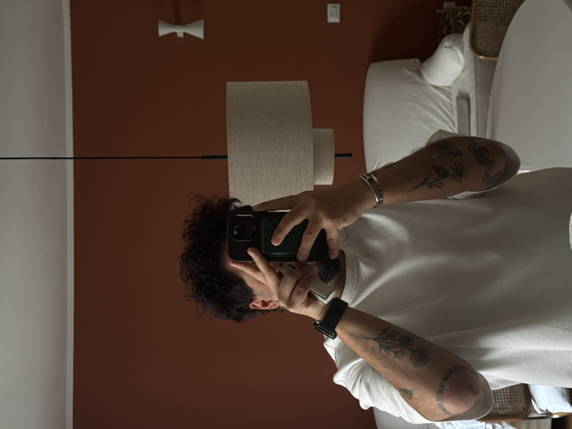 A person with dark, curly hair and fair skin is holding a smartphone and taking a selfie in what appears to be a bedroom or hotel room. The person is wearing a white t-shirt, a black smartwatch, and at least one silver bracelet. Tattoos are visible on both arms.
The background includes a rust-colored wall with a white wall on the left side of the frame. There's a modern light fixture on the rust-colored wall and a smaller white device, potentially an outlet or switch, near the top of the wall. A floor lamp with a neutral-colored shade is also visible. To the right of the person, there is a white bed and a rattan chair.
It's not possible to confirm if the location is in Milan, Italy without additional information.