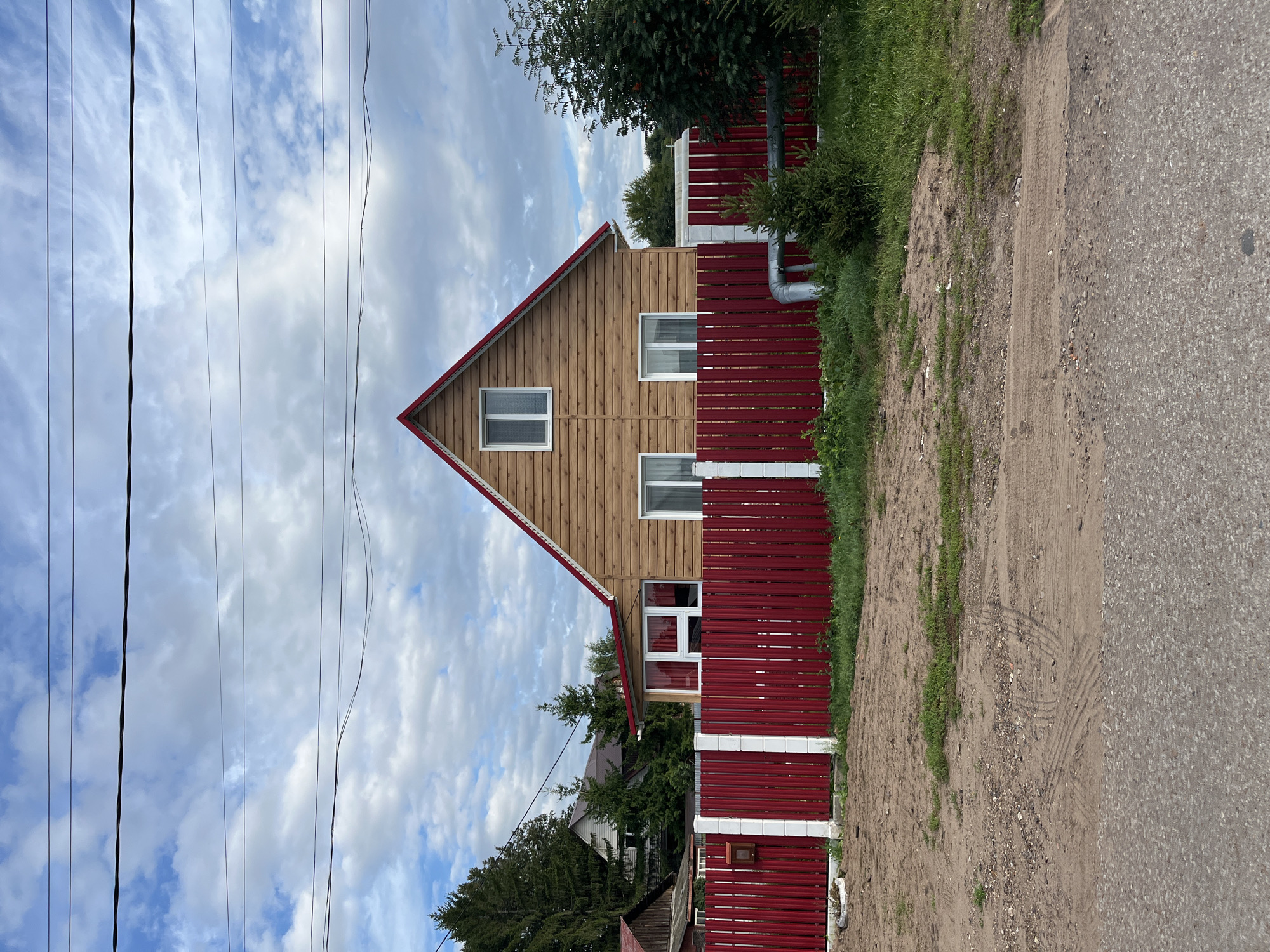 The image depicts a wooden house with a red roof, situated in a rural or suburban area. A bright red vertical slat fence with white posts runs across the foreground. The house has several windows, and appears to be made of horizontal wooden planks. The sky is filled with white, fluffy clouds against a blue background, and there are power lines visible above the house. Trees are present on the left and right sides of the property. The ground in front of the fence is a mix of dirt, gravel, and some sparse green vegetation. A paved surface, likely a road, is visible at the very bottom of the image. The scene appears to be calm and unpopulated, with no people or animals in view. The presence of a mailbox attached to the fence suggests it is a residential dwelling. Based on the architectural style and the natural surroundings, this image could plausibly be from the city of Ufa, Russia, as such houses and fences are common in dacha communities and rural settlements in the region.
