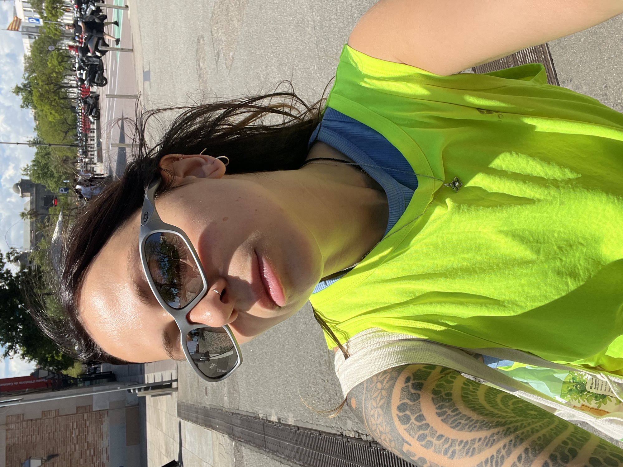 A selfie photograph shows a woman wearing sunglasses and a bright yellow top, outside in an urban environment. She is wearing a nose ring in her left nostril, and the arm visible on the right of the frame is heavily tattooed. The reflection in her sunglasses suggests the presence of trees and buildings across the street. The background shows what appears to be a row of parked motorbikes, with buildings and trees visible further away. Based on visual landmarks such as road markings, railings, and vehicle types, and reflections in the sunglasses, the location is most likely somewhere in the city of Barcelona, Spain.
