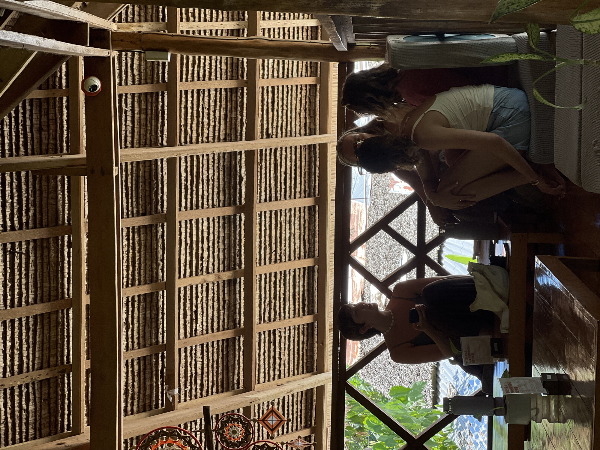 This image depicts an interior scene inside a structure with a thatched roof, possibly a cafe or restaurant, in General Luna, Philippines. Four individuals are present, two women seated on a bench-like structure and two women sitting on the floor. The woman on the left, with short dark hair, is wearing a dark top and holding a phone. Next to her, a woman with blonde hair is looking away from the camera. To the right, a woman in a white tank top and denim shorts is sitting with her legs crossed, and another woman in a red top is seated next to her. Objects on a table in the foreground include a grey thermos, a potted plant with a sign, and a light-colored bag. The background shows a view of lush greenery and a textured surface, possibly a wall or another building. The overall atmosphere suggests a casual, relaxed setting.