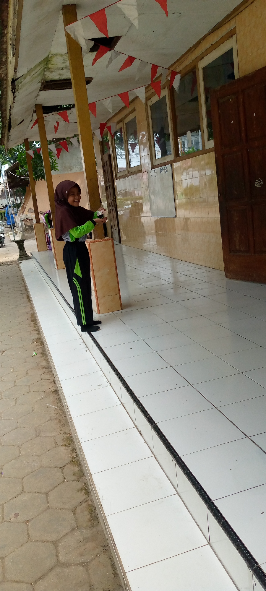 The image shows a young girl wearing a brown hijab and a green and black tracksuit, standing on a tiled outdoor walkway. She is smiling and holding a small white object in her hands. The walkway is bordered by a dark, textured strip. Behind her, there is a building with light-colored walls, several windows, and a wooden door. Red and white triangular flags are strung along the overhang of the building. The ground to the left of the walkway is paved with hexagonal concrete blocks. The scene appears to be in an outdoor area, possibly a school or public building, in Pacing, Indonesia, suggested by the presence of Indonesian flags.