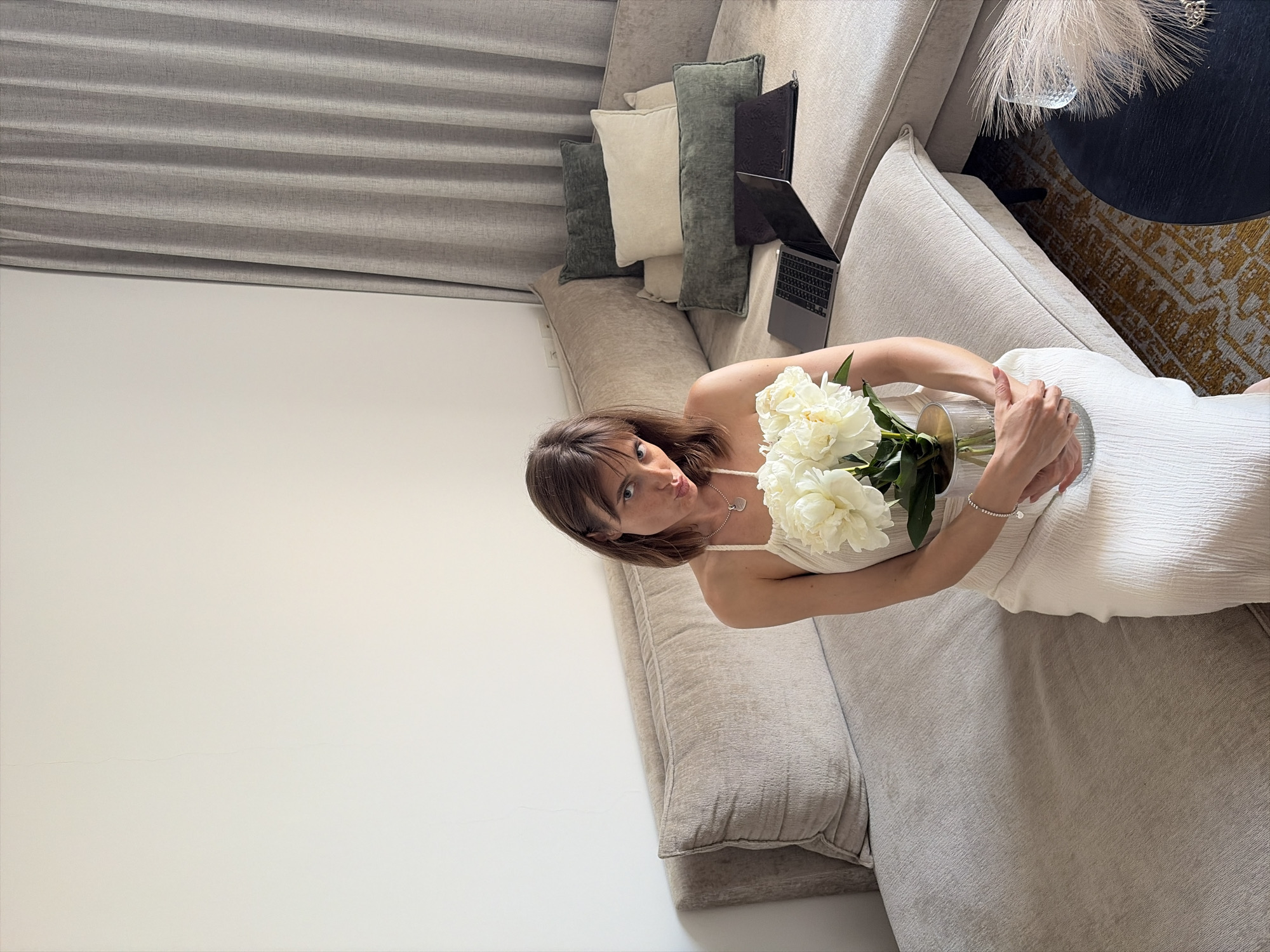 A woman is lying on a light-colored sectional sofa, holding a bouquet of white peonies in a glass vase. She is wearing a white sleeveless dress and a necklace with a heart-shaped pendant. Her right hand rests on the vase, and her left arm is bent, with her hand resting on the sofa. Her facial expression is playful, with her lips puckered as if blowing a kiss. The sofa is adorned with several decorative pillows in varying shades of green and cream. A laptop is open on the sofa next to the pillows, and a dark-colored side table with a decorative light fixture is visible in the background. The room is softly lit, suggesting daytime, and the overall atmosphere appears relaxed and intimate. The woman is positioned in the center of the frame, with the sofa extending to the left and right. The background includes a light-colored wall and a portion of what appears to be blinds on the left side of the image. The image is shot from a slightly elevated angle, looking down on the woman and the sofa. No visible text is present in the image.