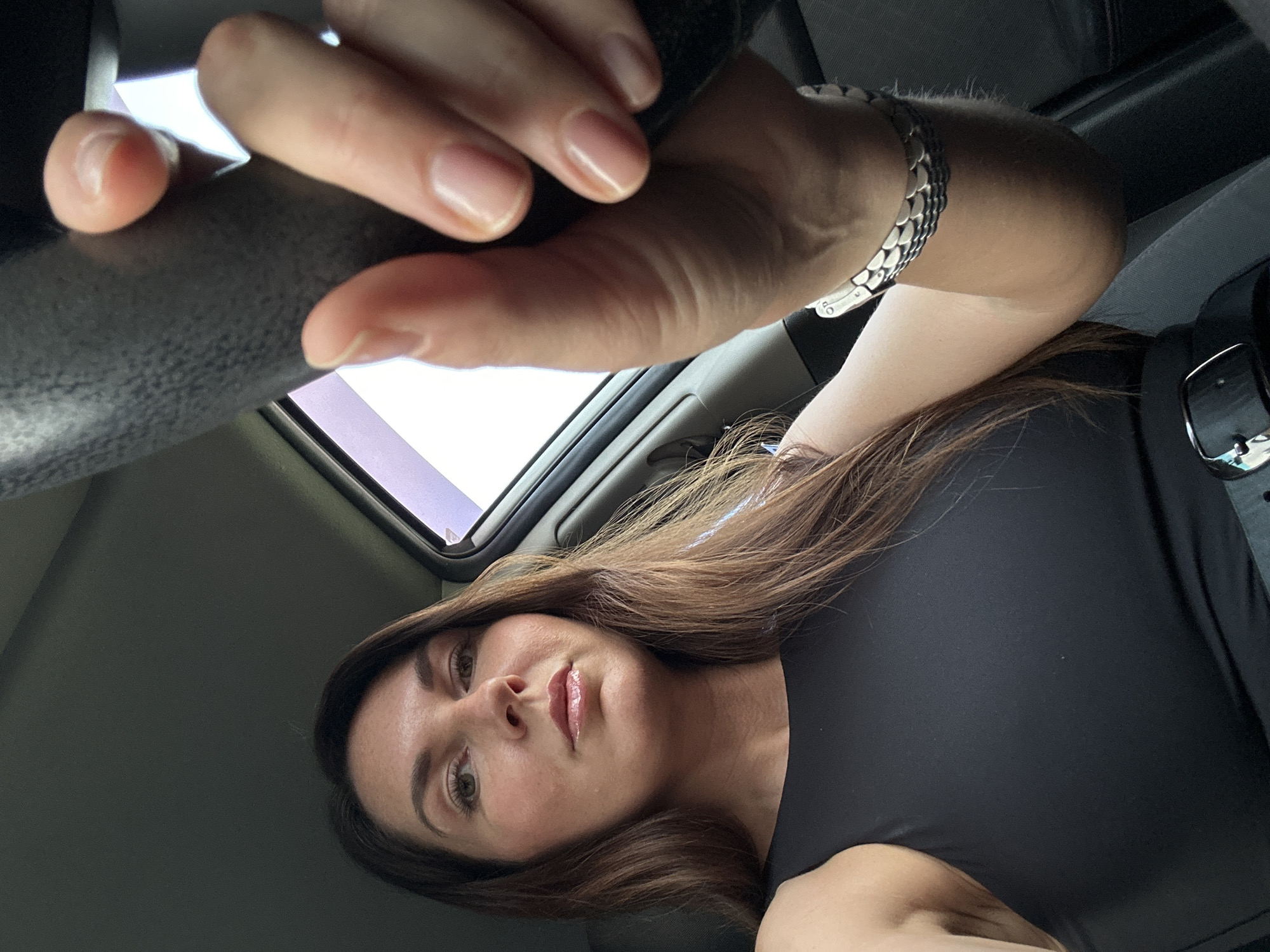 The image shows a woman in the driver's seat of a car, holding the steering wheel. She has long brown hair and is wearing a black sleeveless top. A silver bracelet is visible on her wrist. The interior of the car is dimly lit, with sunlight filtering in from the side window. The background appears to be a blurred outdoor scene, suggesting the car is in motion or parked on a street. The location is not specified in the image itself.