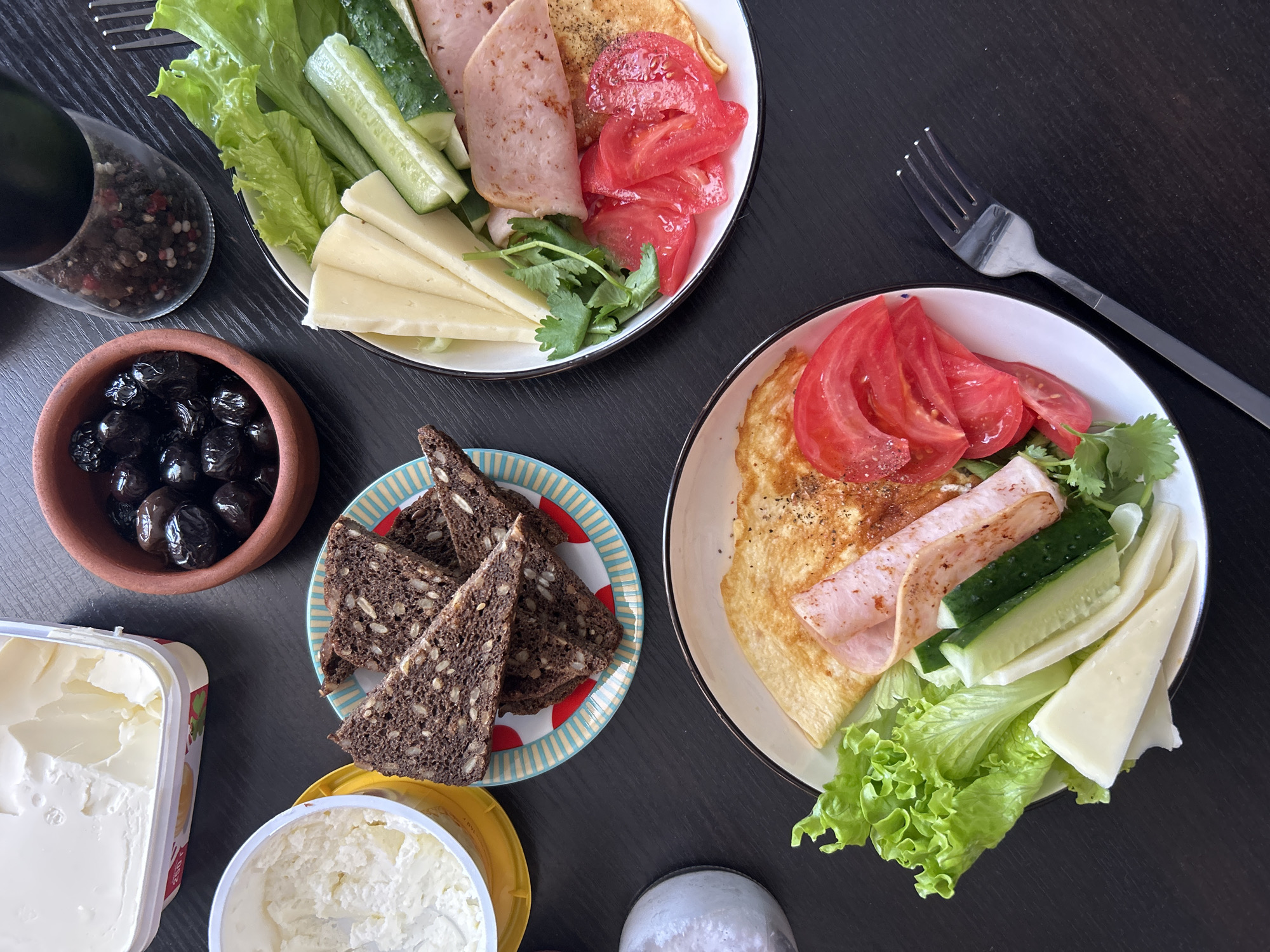 A top-down view shows a table in an unspecified location. Two plates, each containing an omelet, sliced tomatoes, sliced cucumber, ham, cheese slices, and lettuce, are visible. A bowl of olives, a dish with dark rye bread slices, a container of cream cheese, and a pepper grinder are arranged around the plates. Two forks are also on the table. The scene suggests a meal setting.