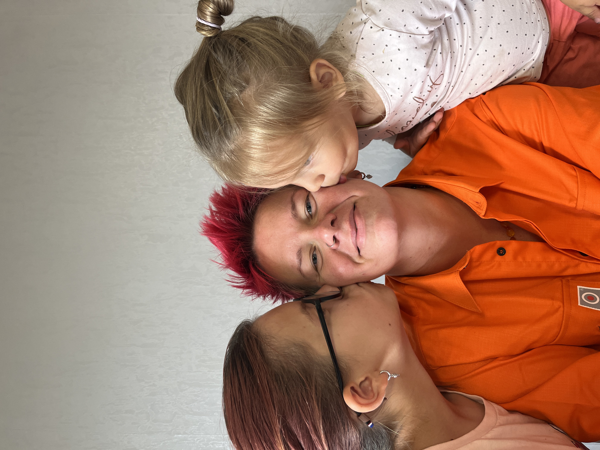In this close-up, waist-up photograph, a person with bright red, spiky hair is being kissed on both cheeks by two children. The person in the center is wearing an orange collared shirt with a logo patch on the left chest. They have a nose ring and appear to be smiling or amused.
On the left, a child with dark hair and glasses is kissing the person's left cheek. This child is wearing a light-colored t-shirt.
On the right, a younger child with blonde hair tied in a bun is kissing the person's right cheek. This child is wearing a white t-shirt with dark polka dots and a pink garment on the lower half of their body.
The background is a plain, textured light gray wall. There are no visible indications of the city of Chaikovsky, Russia, or any specific urban environment. The image is indoors and focuses solely on the interaction between the three individuals.