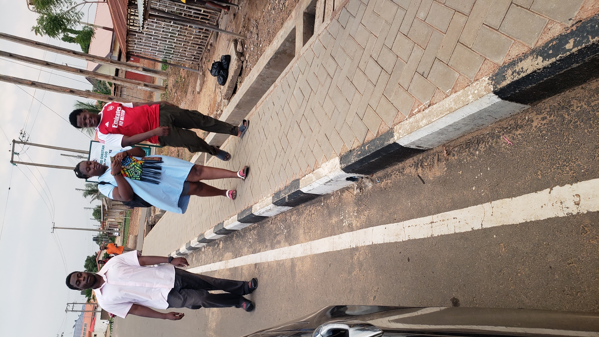 The image shows three people standing on a road next to a sidewalk. The road has a painted white line marking a lane. At the bottom of the frame is a portion of a black vehicle.
The person closest to the camera is wearing a light pink collared shirt and dark pants. The second person is wearing a light blue dress and holding a woven bag. The third person is wearing a red Arsenal jersey and dark pants.
The sidewalk is made of interlocking pavers and has a painted curb with alternating black and white stripes. There are buildings and trees visible in the background.
Based on the provided location of Narayi, Nigeria, the image depicts a scene in that city.