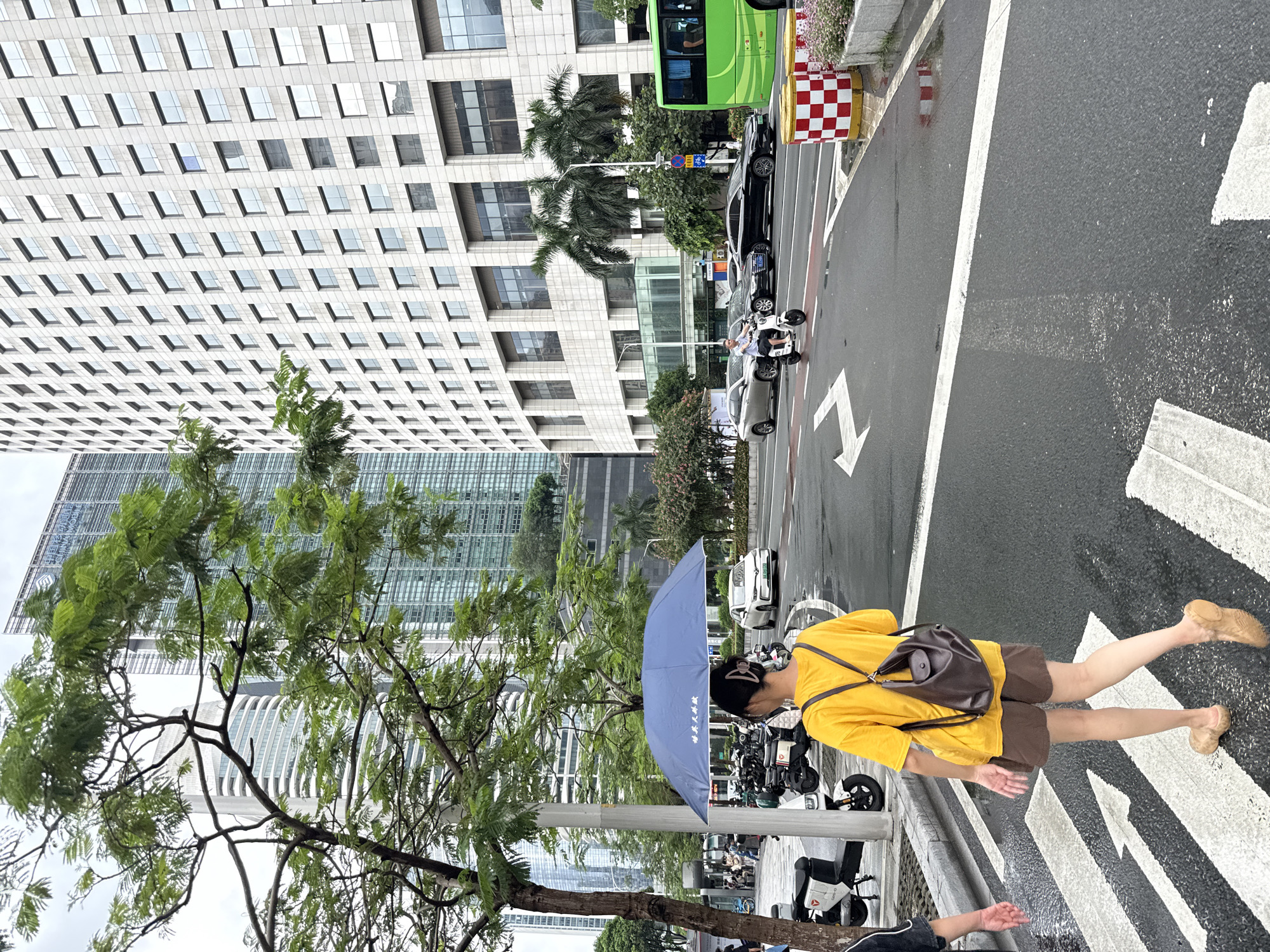 The image captures a street scene in Tianhe District, China, with a woman walking across a crosswalk while holding an open blue umbrella. She is wearing a yellow t-shirt and brown shorts, with a dark brown backpack slung over her shoulders. In the background, modern high-rise buildings dominate the skyline, featuring numerous windows. To the right of the woman, a green bus is visible. Several parked scooters and a white electric scooter with a rider are also present on the street. The road appears wet, suggesting recent rain. Traffic lines and white pedestrian crossing markings are clearly visible on the asphalt.