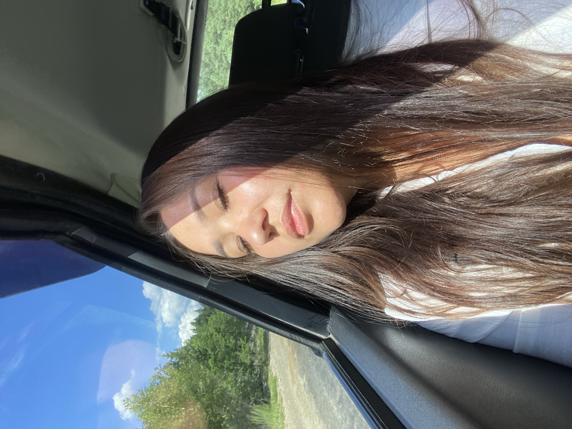 In Zilair, Russia, a young woman with long, dark hair sits inside a vehicle, her eyes closed as sunlight streams through the window, casting a warm glow on her face. She wears a white shirt. Outside the car, a dirt road winds through a lush, green forest under a bright blue sky dotted with fluffy white clouds. The scene captures a serene moment during a drive through the natural landscape surrounding Zilair.