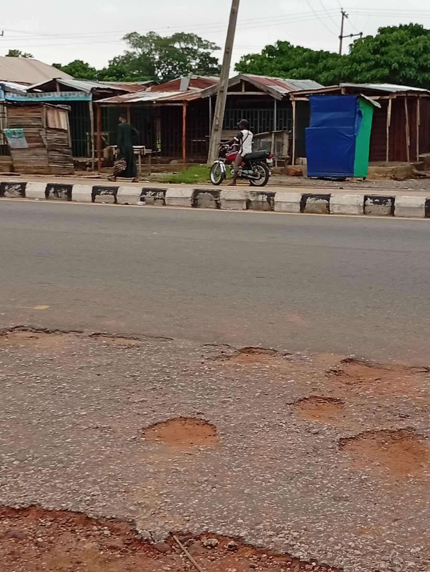 The image depicts a street scene in Lokoja, Nigeria. In the foreground, the asphalt road surface is damaged with potholes. Beyond the road is a sidewalk marked with black and white painted curbs. Along the sidewalk are several small, makeshift shops or stalls constructed of wood and corrugated metal.
A man in a dark green robe is walking past one of the shops, carrying a bag. A motorcyclist is standing next to a red and black motorcycle, positioned in front of another shop draped with blue and green tarps. The overall scene appears to be a typical street scene in a developing urban environment. The sky is overcast. Green foliage is visible in the background, suggesting a tropical climate.