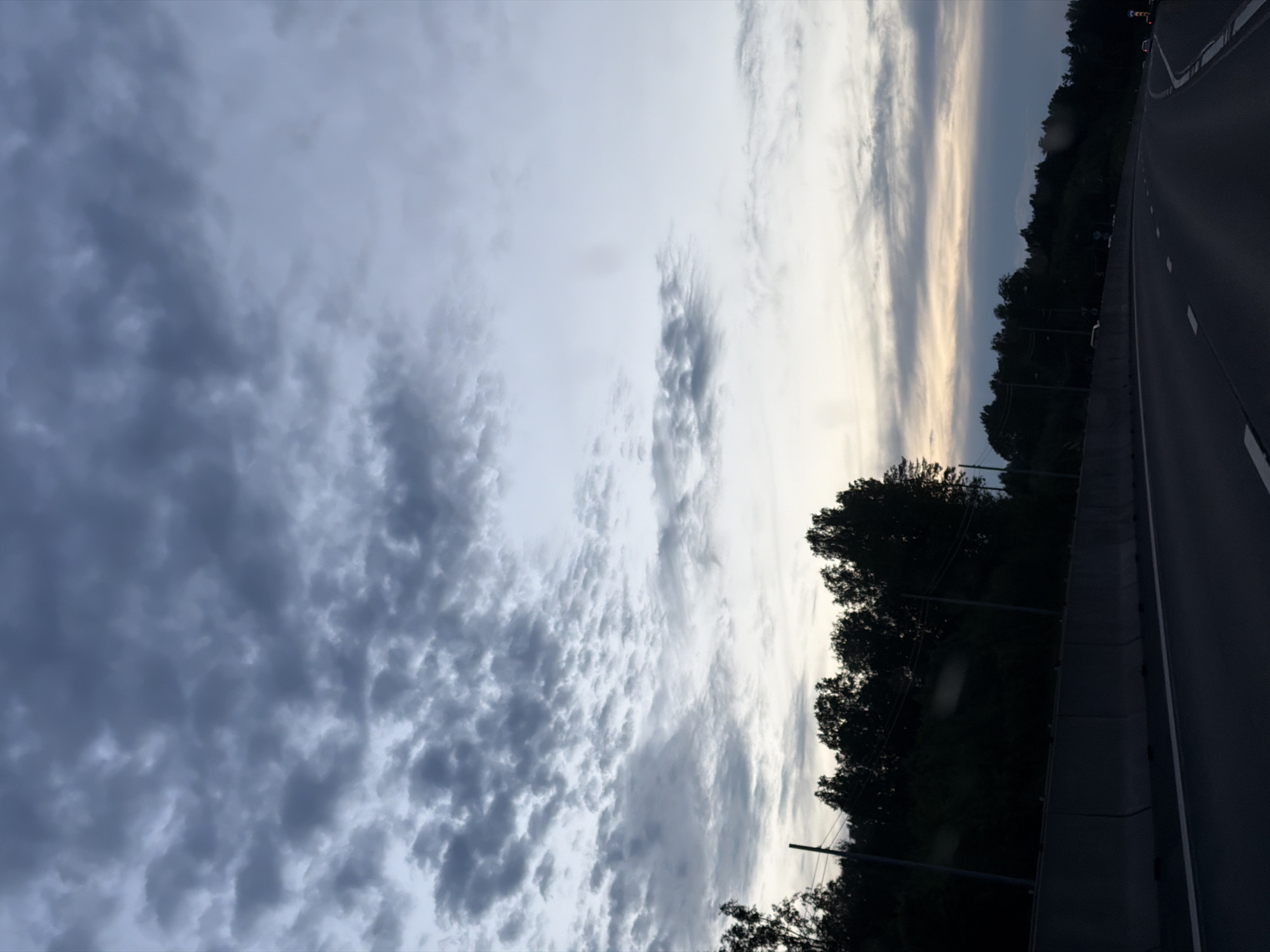 This image, taken in Kerro Massiv, Russia, captures a dramatic highway scene at dusk or early evening. The perspective appears to be from a vehicle on the road, looking towards the right side of the highway and a vast, cloud-filled sky.
The sky dominates the upper and left portions of the frame, showcasing a diverse cloudscape. On the left, dense, dark grey, textured clouds create a heavy, brooding atmosphere. As the eye moves right and upwards, the clouds become lighter, transitioning into streaky white and pale yellow formations, indicating the presence of the setting sun, which casts soft light on the horizon.
On the right side of the image, a multi-lane asphalt highway is visible, gently curving into the distance. It is marked with clear white dashed and solid lines. A robust grey concrete barrier separates the highway from a dense line of dark, silhouetted trees that border the road. A few dark poles, possibly utility poles or streetlights, can be seen within the treeline and alongside the barrier. Faint lights from distant vehicles are discernible on the highway. No visible text is present in the image. The scene conveys a sense of travel amidst natural elements under a dynamic, cloudy sky.