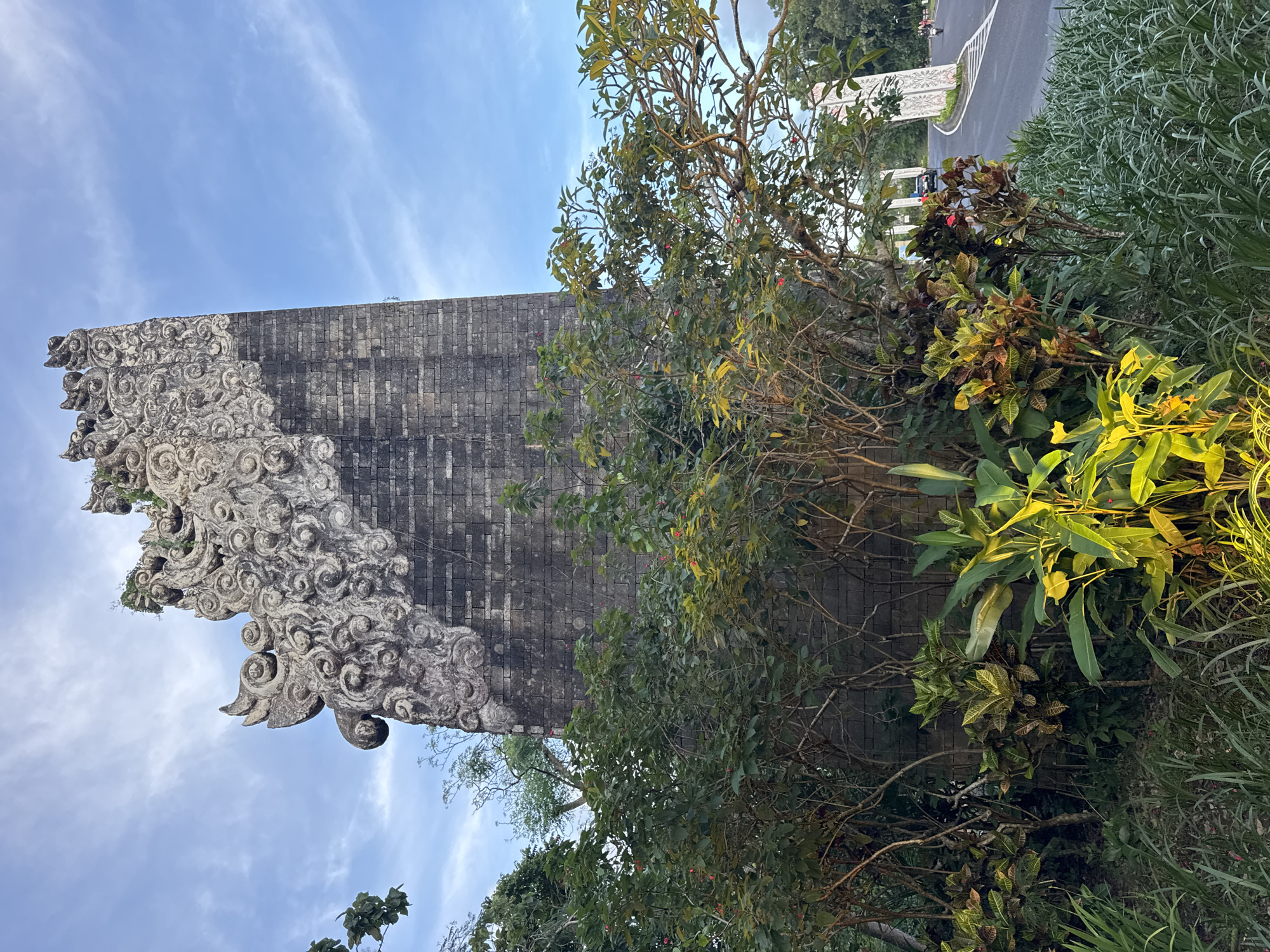 This is a medium shot taken at eye-level, showcasing an ornamental brick gate or wall in Kuta Selatan, Indonesia. The top of the wall has intricate, stylized carvings. It is adjacent to a street with a curb visible in the background. The wall is partially obscured by a variety of tropical shrubbery and trees. The sky above is blue, with light, wispy clouds visible.