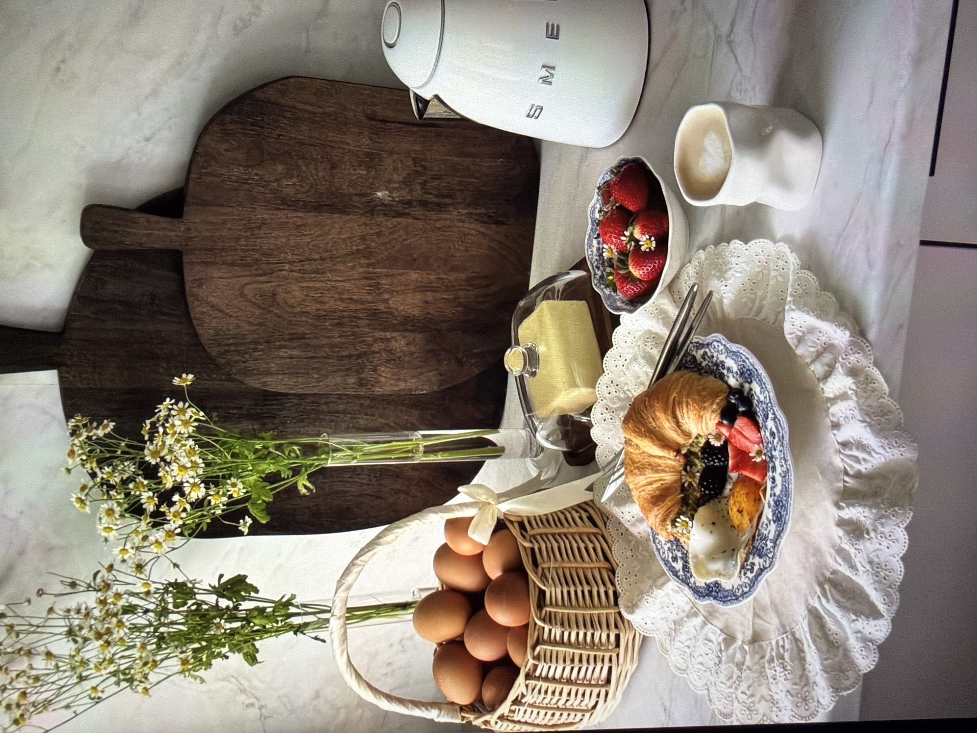 The image displays an inviting, overhead view of a breakfast or brunch setup, carefully arranged on a bright, possibly marble-patterned countertop, likely indoors in Ros Na Rí, Ireland. The scene suggests a styled flat lay, possibly for a photoshoot.
Key subjects include kitchenware and fresh food items. A white SMEG electric kettle is prominently placed alongside two dark wooden chopping boards of differing sizes. A clear glass vase holds a bouquet of fresh white and yellow chamomile flowers. A rustic wicker basket, tied with a cream ribbon, overflows with numerous brown eggs.
Central to the food display are a white ceramic bowl filled with fresh red strawberries, some garnished with small white flowers, and a white, organically shaped ceramic mug containing a hot beverage with visible latte art. A blue and white patterned plate, resting on a white lace-trimmed placemat, holds a golden croissant, a vibrant mix of fresh berries (blueberries, blackberries, and strawberries), and a single egg. A pair of metal chopsticks lies across the plate. A small glass cloche covers a yellow block, likely butter or cheese.
The arrangement evokes a serene and pleasant morning atmosphere, enhanced by the fresh flowers and natural light, indicating it is daytime. The visible text "SMEG" is clearly seen in silver letters on the electric kettle.