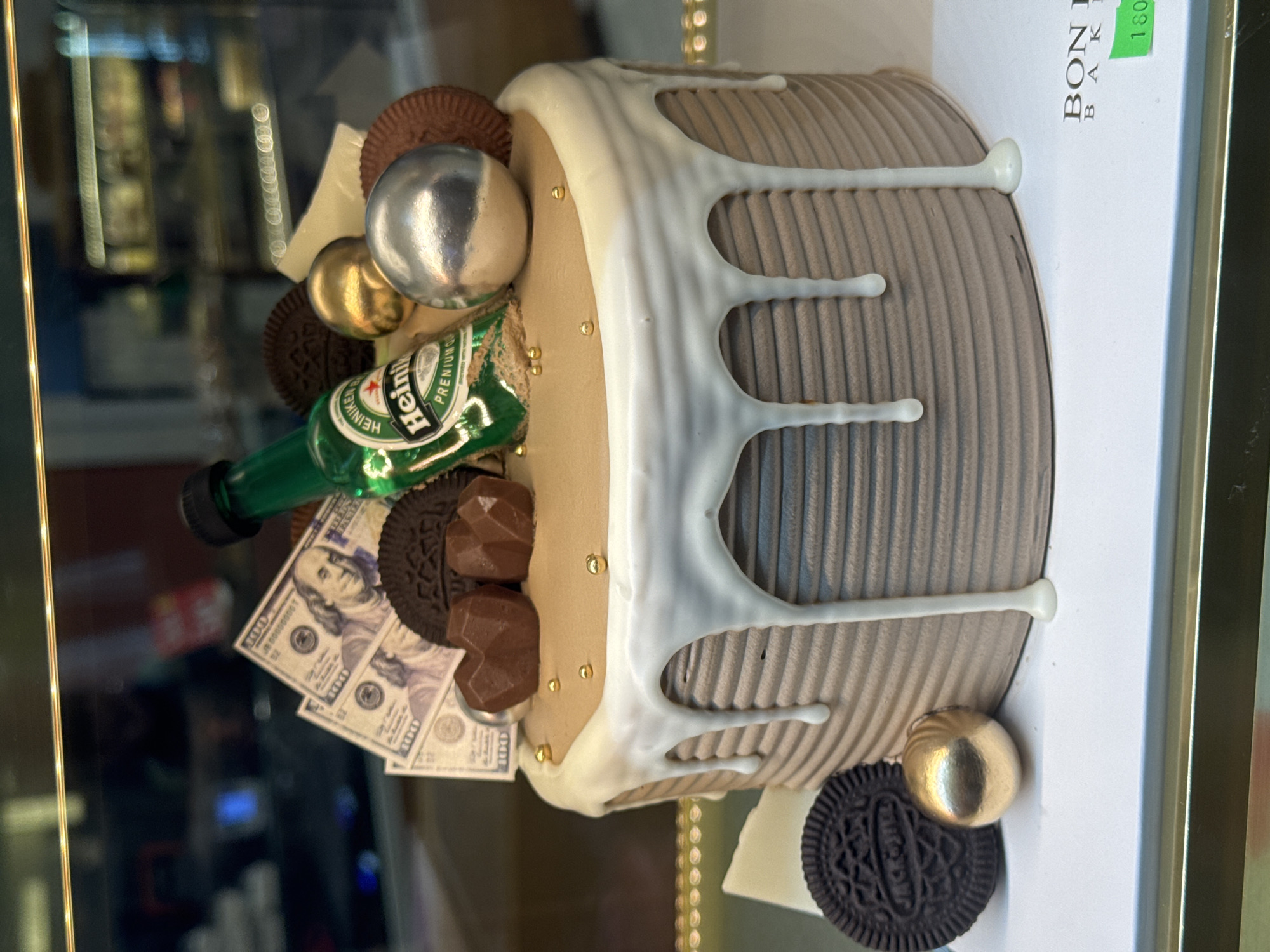 A chocolate cake is displayed in a bakery case. The cake is decorated with a white drip glaze, chocolate cookie crumbles, chocolate candies, gold and silver spheres, fake dollar bills, and a miniature bottle of Heineken. The cake sits on a white board. A label with the words "BON BAKE" and the number "180" is visible on the board. The background is blurred and shows shelves with baked goods. The overall impression is a themed cake designed for an adult celebration, possibly a birthday or an accomplishment. The location is a bakery in Da Lat, Vietnam.