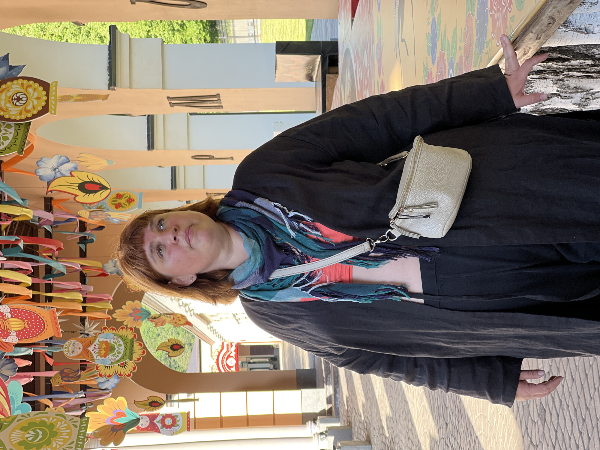 A fair-skinned woman with reddish-brown hair and bangs stands facing slightly left while looking upwards and to her right. She wears a dark, long-sleeved open cardigan over a coral crop top, dark pants, and a multi-colored fringed scarf. A light beige textured crossbody bag hangs from her right shoulder. Her left hand is by her side, and her right hand rests on a low, patterned surface to her right. The scene is set in Estosadok, Russia.
Above her, numerous colorful, flat, cut-out decorations depicting Russian folk art motifs such as matryoshka dolls, stylized flowers, and birds hang from an overhead structure, intertwined with various colored ribbons. She is positioned under an architectural colonnade featuring light peach-colored walls and white arched pillars. Some pillars display dark, decorative letter-like elements. Beyond the arches, a building with red architectural details and lush green foliage are visible in the background. The ground is paved with patterned light-colored blocks. Strong sunlight casts distinct shadows across the pavement and structures, indicating bright outdoor conditions. To her immediate right, a section of a light-barked tree trunk is visible next to the patterned surface.