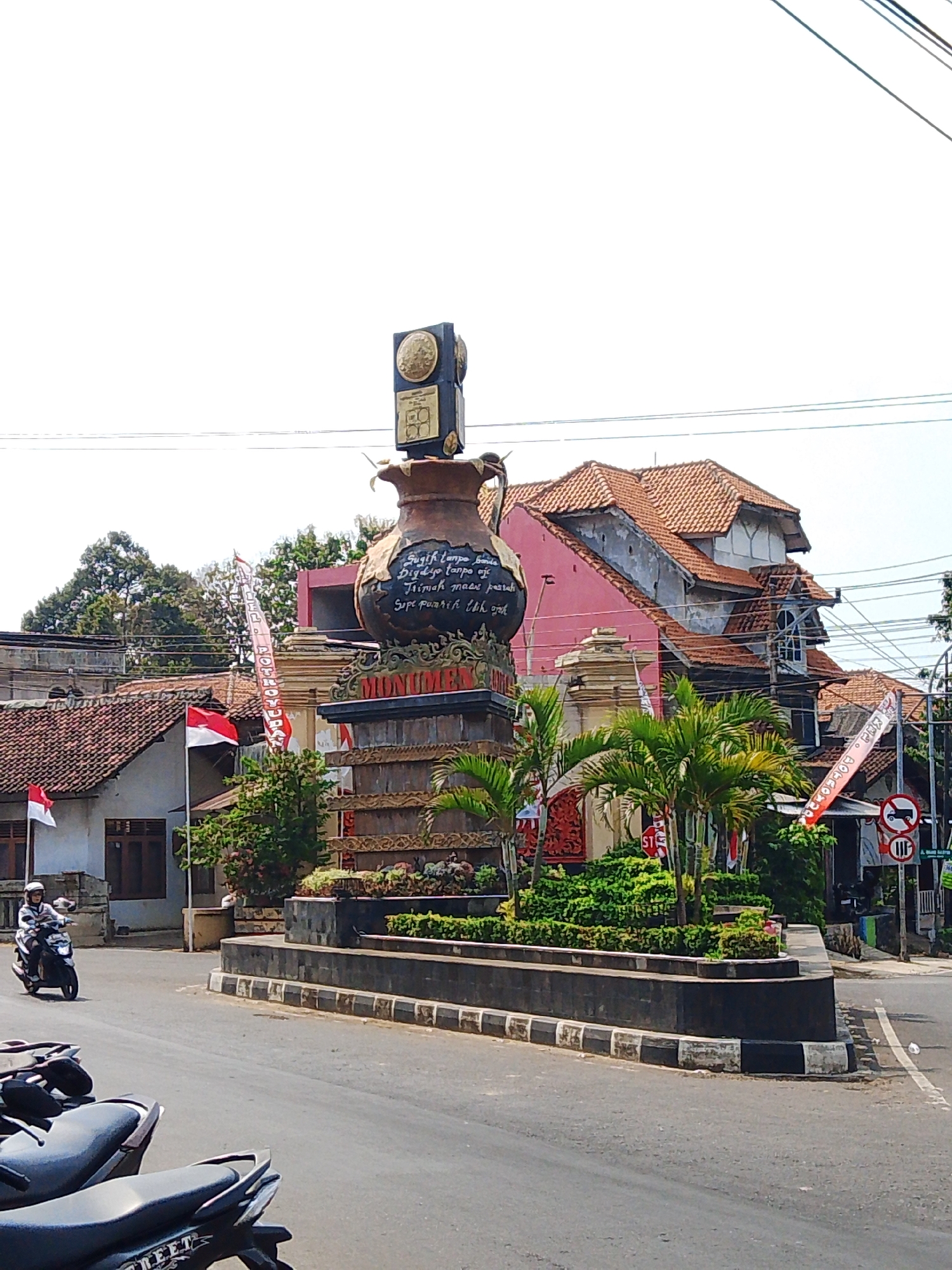 In Potroyudan, Indonesia, a monument stands at a street intersection, featuring a large decorative jug atop a tiered base adorned with gold accents. A rider on a scooter travels past the monument. The area is surrounded by buildings with tiled roofs and trees, and several Indonesian flags are displayed. Power lines are visible overhead.