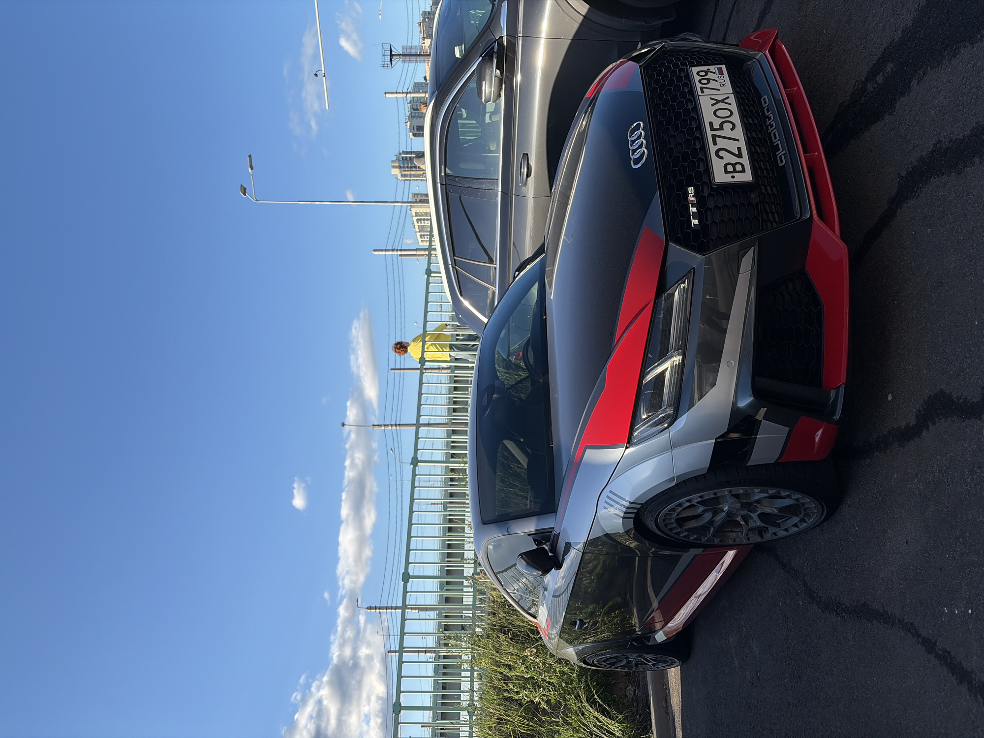 An Audi TT RS sports car, with a black, silver, and red custom paint job, is parked on asphalt. The car has a Russian license plate reading "B275OX 799". In the background, a dark gray SUV is parked next to it. Behind the cars, a green metal fence separates the parking area from a raised structure, possibly a railway bridge, with power lines. A person in a bright yellow jacket stands on the other side of the fence, looking towards the right. In the distance, several industrial buildings and smokestacks are visible against a blue sky with scattered white clouds. The scene appears to be in an industrial or commercial area within Saint Petersburg, Russia, given the license plate and the architectural style of the distant buildings.