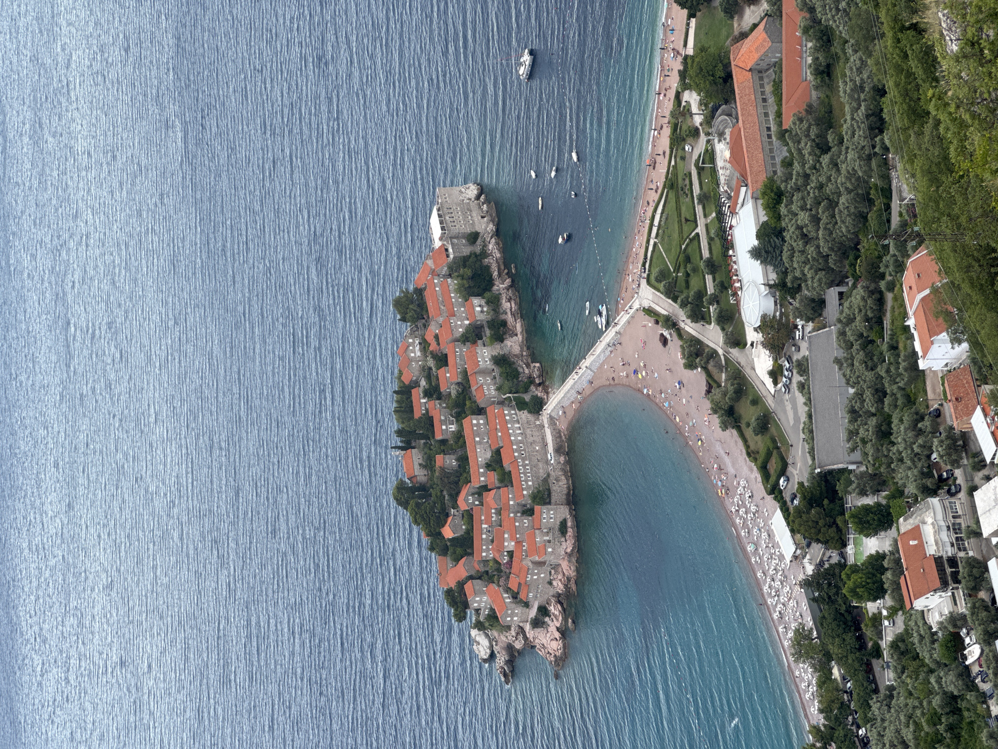 This is an aerial, top-down photograph of Sveti Stefan, Montenegro. The image showcases the iconic islet, a former fishing village now a luxury resort, connected to the mainland by a narrow causeway. The islet itself is densely packed with traditional stone buildings featuring terracotta roofs. Surrounding the islet is the calm, blue-green Adriatic Sea, showing ripples from gentle movement.
On the mainland, a crescent-shaped sandy beach is visible, lined with numerous beach chairs and umbrellas, indicating a populated beach. People are scattered across the sand, enjoying the sunny weather. Adjacent to the beach, there are well-maintained green spaces with trees and pathways, leading to several buildings, including hotels and villas, also with tiled roofs. The presence of parked cars suggests visitor accessibility.
The scene conveys a sense of a popular tourist destination on a clear day, likely during the warmer months. The lighting suggests it could be late morning or early afternoon. The overall mood is one of leisure and vacation. No visible text is present in the image.