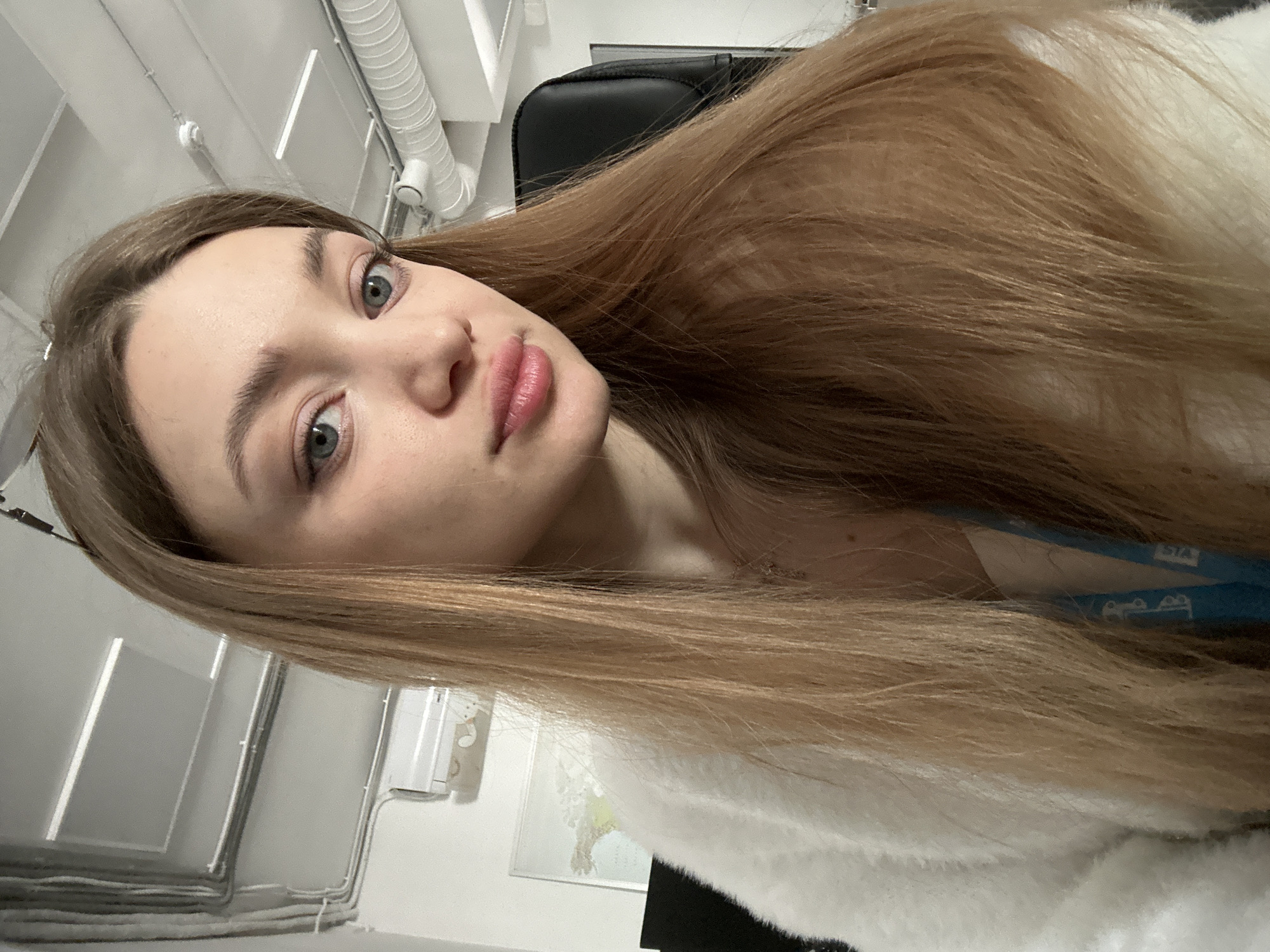 An indoor, diagonally oriented shot features a young woman with long, light brown hair looking directly at the camera. Her hair, appearing light blonde in sections, flows over her shoulders and arm. She has blue eyes, light eyebrows, and lightly made-up pink lips. Her expression is neutral to slightly serious. She is wearing a white, fluffy garment, possibly a sweater or jacket. A light blue strap or lanyard with white, indistinct text or a logo, including a stylized "SA" and two smaller rounded shapes below it, is visible near her neck.
The background suggests an office or commercial setting. Overhead, a white ceiling with several large, white cylindrical ventilation ducts and rectangular light fixtures is visible. Behind her, part of a black, upholstered office chair can be seen. To her left, a white wall features some exposed wiring or cables running horizontally. A small, framed map or picture is affixed to this wall, along with a white, box-like object, possibly a small electronic device. The lighting is artificial, casting no strong shadows, indicating an indoor environment without natural light cues. The overall scene is clean and modern.