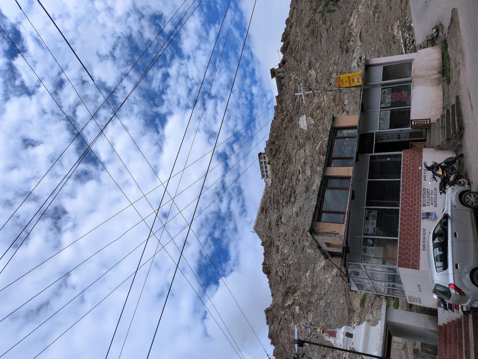 The image depicts a scene in Leh, India, focusing on a roadside establishment set against a rocky hillside. Dominating the upper portion of the frame are numerous power lines crisscrossing against a blue sky filled with white clouds. In the mid-ground, a two-story building appears to be a cafe or restaurant, with a glass-fronted ground floor and a brick facade. Signs on the building indicate "BIKE FOR RENT" and "MOUNTAIN DELIGHT ROOF TOP RESTAURANT". A yellow sign with text reads "OH-LALA CAFE & SANDWICH BAR". In front of the building, a silver hatchback car is parked, and two motorcycles are also visible. To the left of the building, a white Buddhist stupa stands next to a staircase. The rocky slope behind the building is dotted with structures, suggesting buildings or monasteries situated on higher ground. The overall environment suggests a commercial area on the outskirts of Leh, with a blend of local architecture and modern amenities.