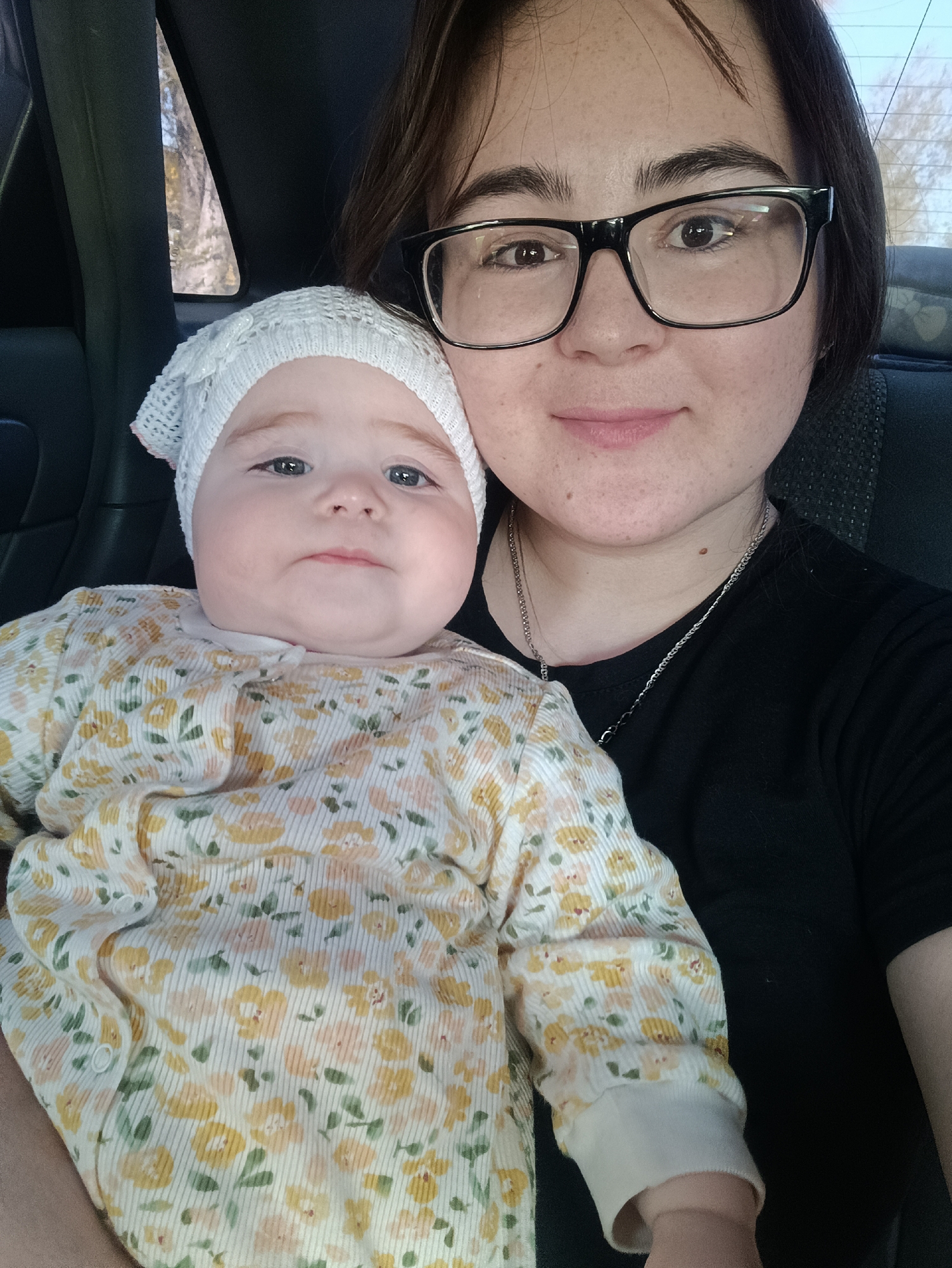 A woman with dark hair and glasses is holding a baby in a car. The woman is wearing a black shirt and a silver necklace. The baby is wearing a white hat and a floral-patterned outfit. The photo appears to have been taken inside a vehicle, possibly in transit. The exact location in Chaikovsky, Russia, cannot be determined from the image.
