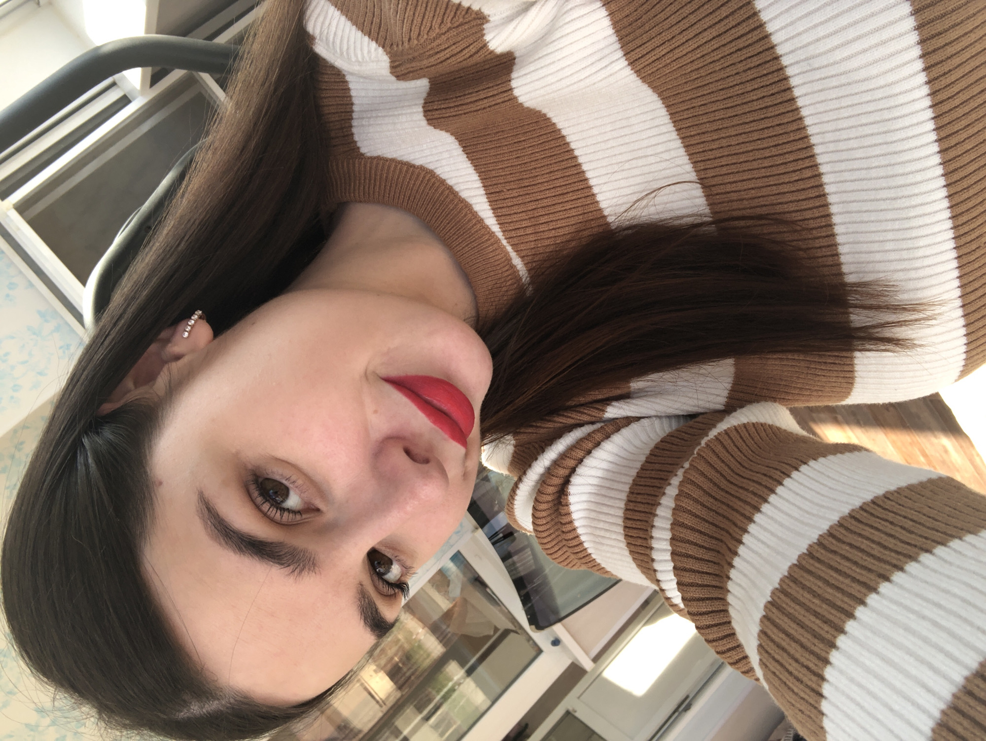 A portrait-oriented photo, rotated 90 degrees clockwise, captures a woman taking a selfie. She has long, dark brown hair that falls over her shoulder, brown eyes, and is wearing bright red lipstick. Her eyebrows are neatly shaped. She wears a brown and white striped, ribbed knit sweater. A small earring, featuring a line of tiny white stones, is visible in her left earlobe.
The background is somewhat blurred but shows reflections of an indoor or outdoor setting through what appears to be a window or glass door. Elements like a dark metal bar, possibly from a chair or railing, and light-colored wall or window frames are visible. The overall lighting suggests it is daytime. The woman has a neutral to slightly smiling expression. There is no visible text in the image. The location is Noviy, Russia, though no specific visual cues confirm this within the image itself.