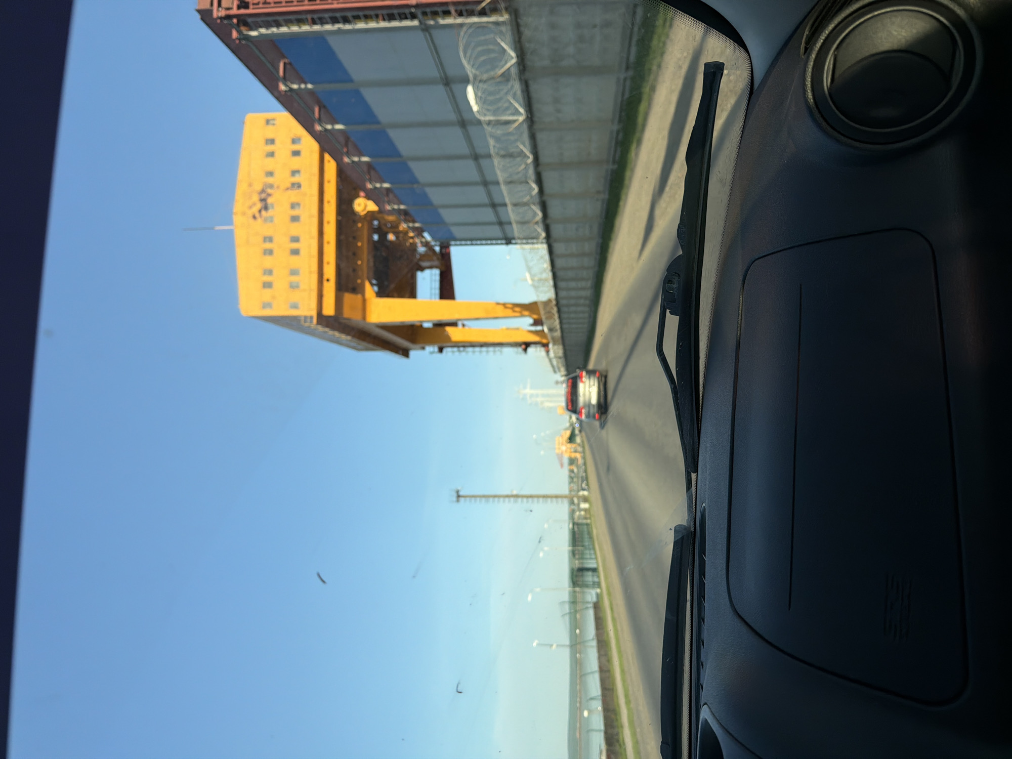 The image, taken from inside a vehicle, shows a road in the city of Chaikovsky, Russia. Visible through the windshield are industrial structures, including a large yellow crane and a building topped with barbed wire, suggesting a secure area. In the distance, other vehicles and infrastructure elements such as light poles can be seen along the road. The sky is clear and blue, indicating daytime. The vehicle's dashboard and windshield wipers are visible in the foreground.