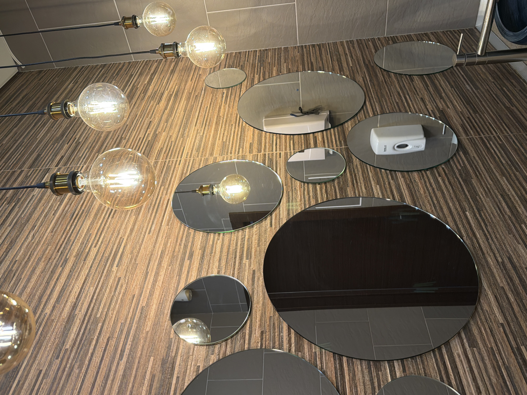 The image displays an interior space, likely a washroom or public restroom, dominated by a wall covered in narrow, horizontally oriented strips of wood-grain material in varied shades of brown. Five spherical, clear-glass Edison-style pendant light bulbs with visible filaments hang at differing heights from dark cords with brass-colored fixtures, emitting a warm yellow light.
Eleven irregularly shaped and circular mirrors of various sizes are affixed to the textured wall. Reflections within these mirrors reveal additional environmental details: a dark brown wooden door, sections of a grey-tiled wall, and a white, wall-mounted soap dispenser labeled "sani.com" mounted on a grey tiled surface. A chrome-plated faucet or water fixture base is partially visible in the bottom right corner, suggesting the presence of a sink out of frame. No individuals or discernible actions are depicted. The specific location within Izhevsk, Russia, is not identifiable from the image.