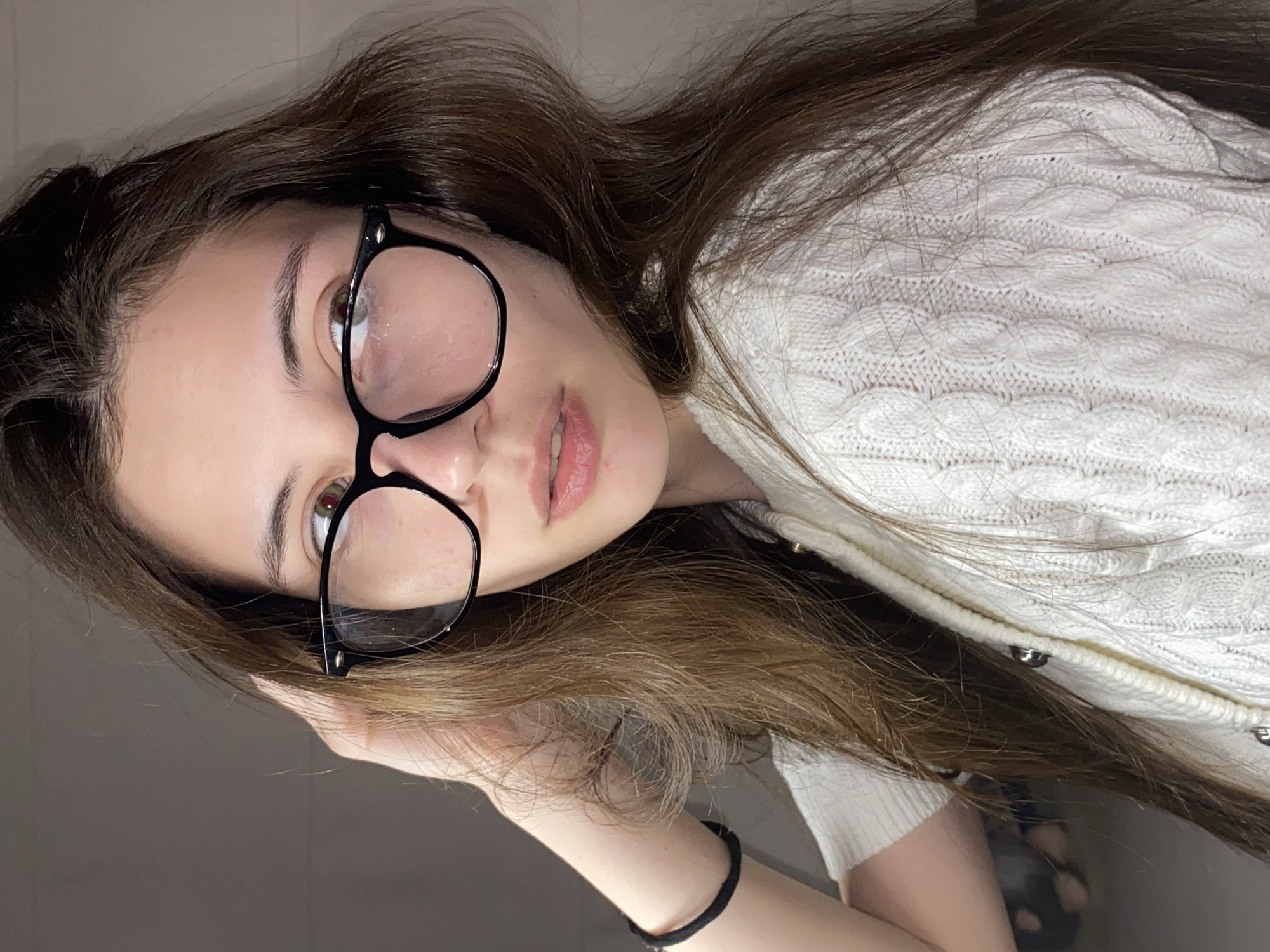 A close-up, vertically oriented portrait features a young woman in an indoor setting, presumably in Votkinsk, Russia. She has long, wavy dark brown hair and fair skin. Her head is tilted, and she looks directly at the camera through black-rimmed glasses. Her eyes appear to be a green or hazel color, with a noticeable reddish tint near the pupil, possibly a reflection from lighting. Her lips are slightly parted.
She is wearing a white cable-knit cardigan with a distinct textured pattern, featuring visible silver-toned buttons down the front opening. Her left hand is seen resting near her hair, and a black elastic hair tie is worn on her left wrist. The background is indistinct and dark, suggesting a plain wall or a dimly lit area, which serves to keep the focus entirely on the subject. The lighting appears to be artificial, casting a soft glow on her face. Her expression is neutral to gently contemplative, conveying a relaxed and personal moment. No text is visible in the image.