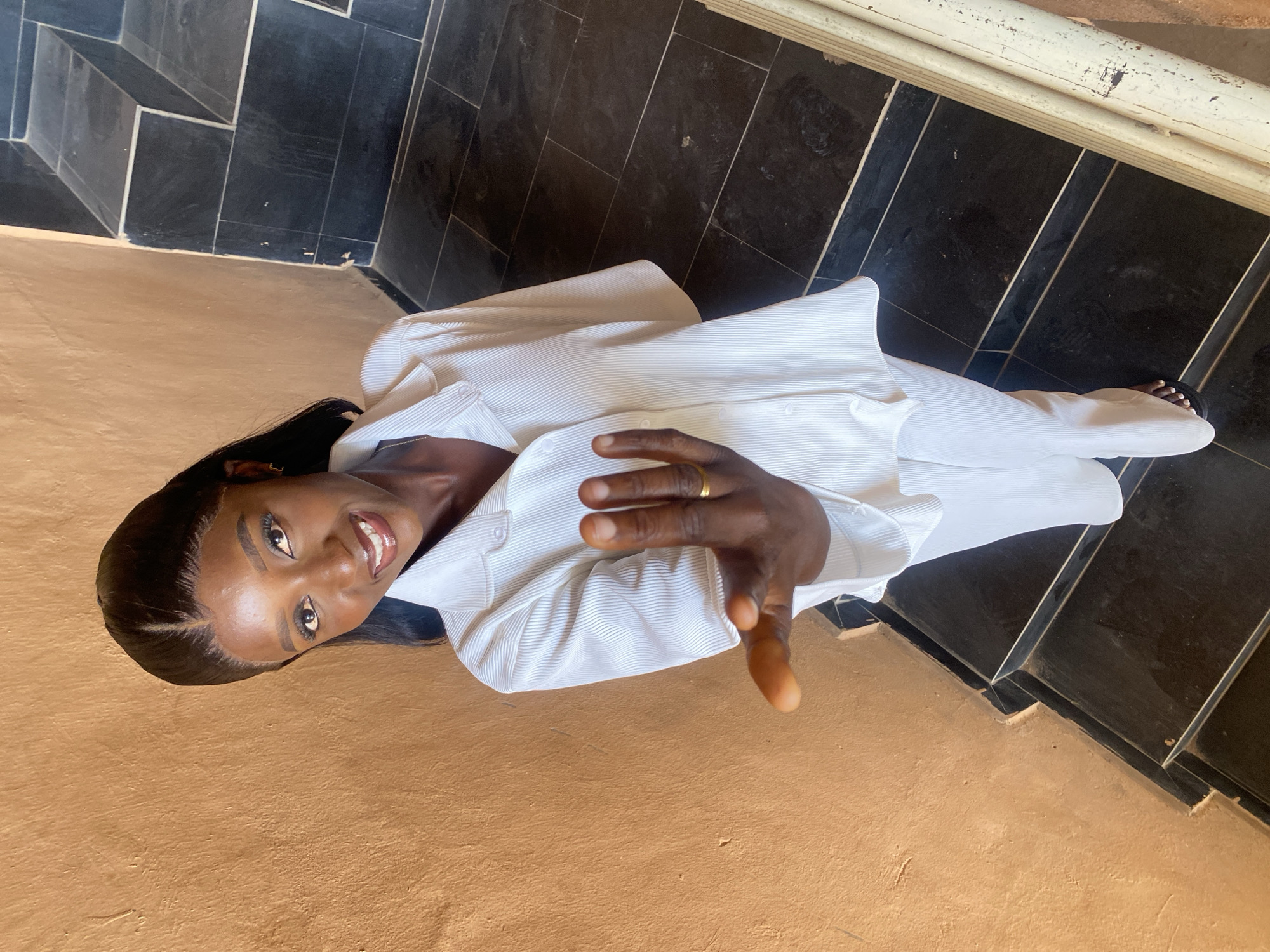 A high-angle, full-body shot shows a young Black woman standing on a staircase in Gura Top, Nigeria. She is looking at the camera and smiling, with her right hand extended towards the viewer. The woman is wearing a white pleated blouse and matching pants. The staircase is made of dark tiles with lighter grout lines, and the wall to the left is a rough, textured, tan color. The lighting suggests an outdoor or semi-outdoor setting with some sunlight present.