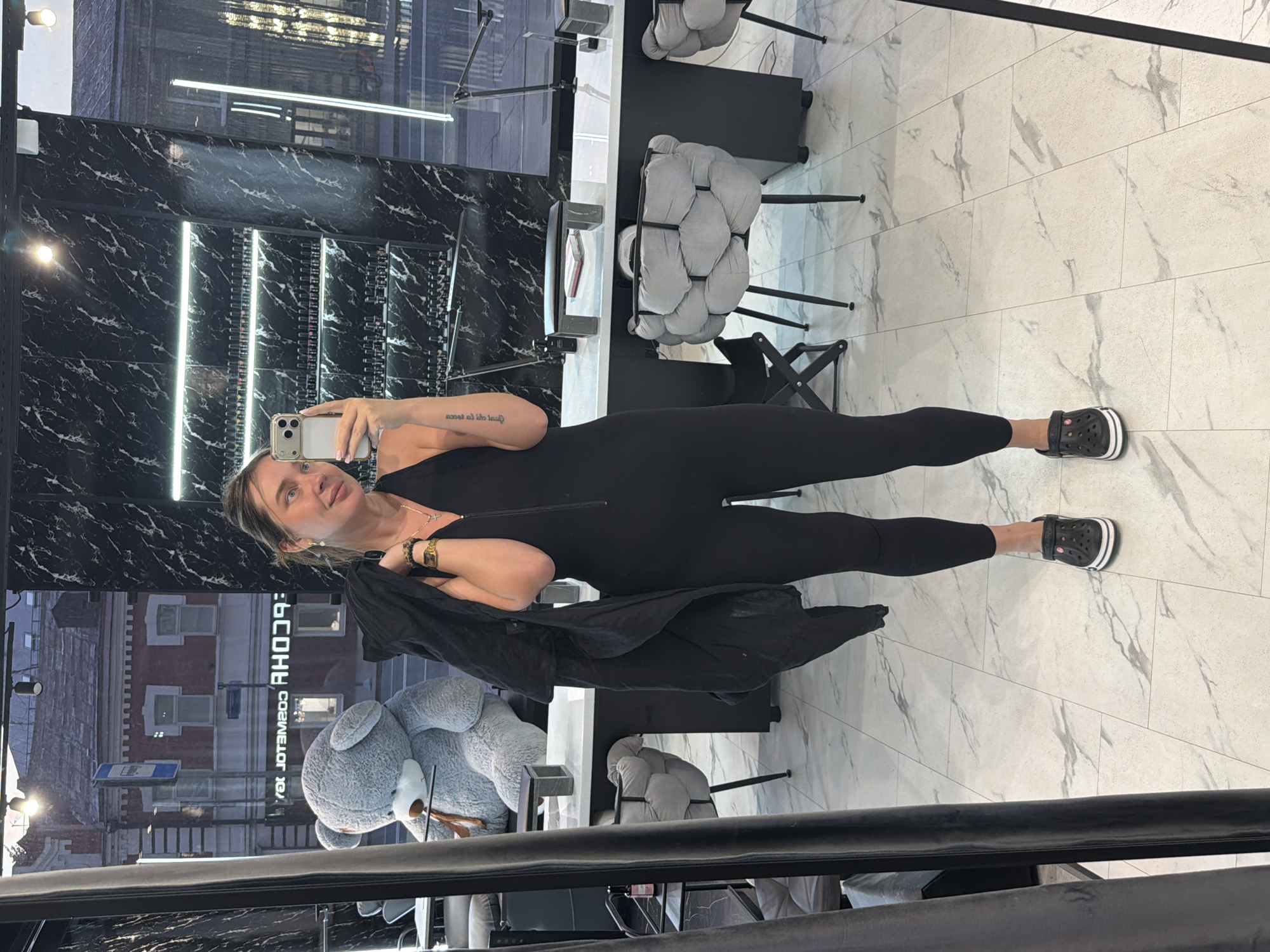 A woman stands in front of a mirror in what appears to be a nail salon. She is wearing a black, sleeveless jumpsuit with a zipper down the front, and black Crocs. She is holding a black jacket over her left shoulder. In the reflection of the mirror, a large gray teddy bear is visible to her left, and beyond that, a street scene with buildings. The floor is tiled with a marble-like pattern. The walls of the salon are decorated with black marble and shelves stocked with bottles, likely nail polish. The scene is lit by overhead lights and strip lighting on the walls. The woman is looking at her phone, which she is holding up to take a selfie.