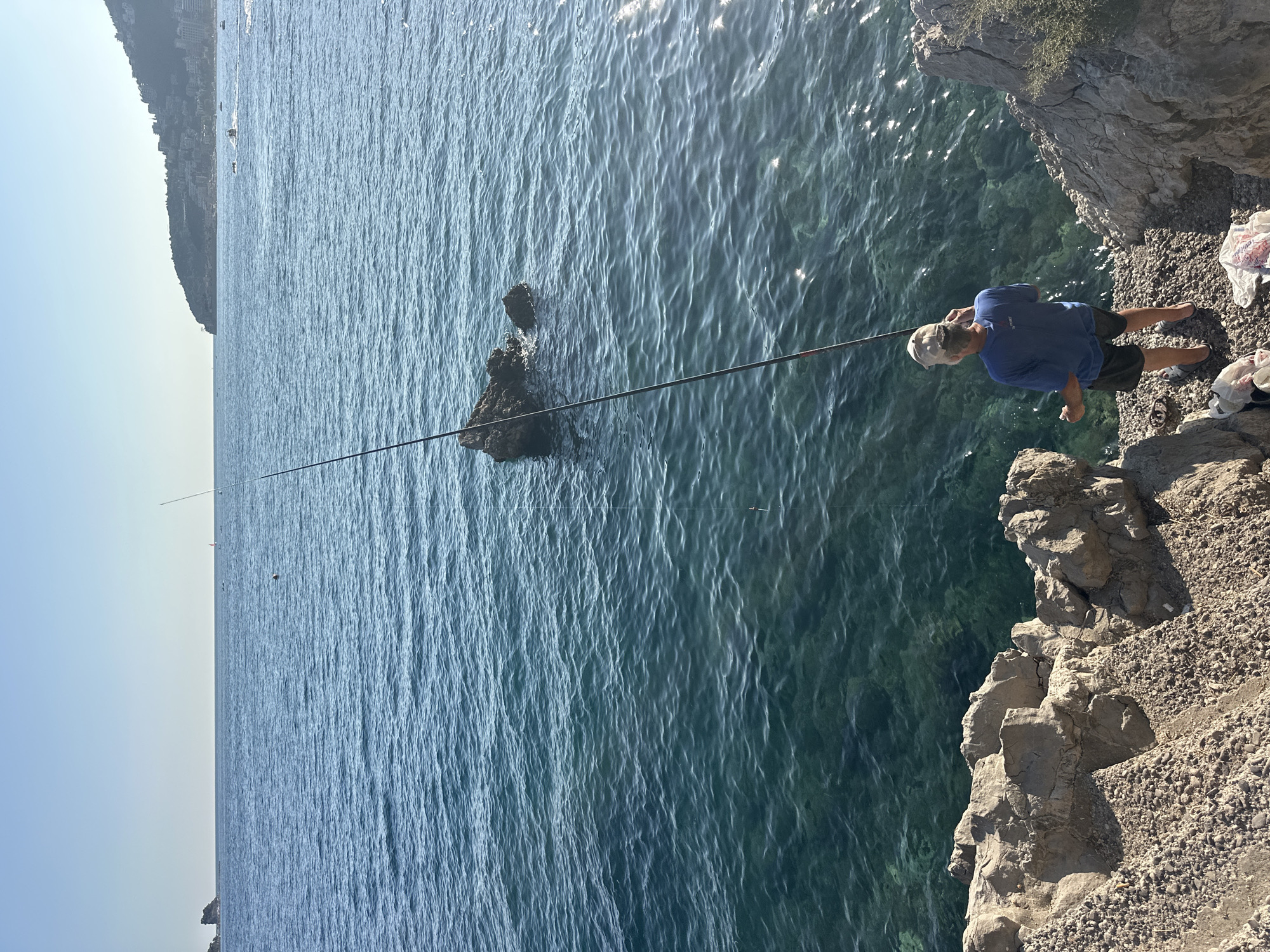 A lone fisherman, wearing a blue t-shirt, dark shorts, and a light-colored baseball cap, stands on a rocky shoreline in Budva, Montenegro, holding a fishing rod. The water is a clear turquoise, with ripples and sunlight glinting off the surface. In the distance, islands and a coastline with buildings can be seen under a clear blue sky. The fisherman is positioned to cast his line into the sea. Several plastic bags are scattered on the ground near his feet. The rocks in the foreground are rough and varied in texture, with a scattering of pebbles.