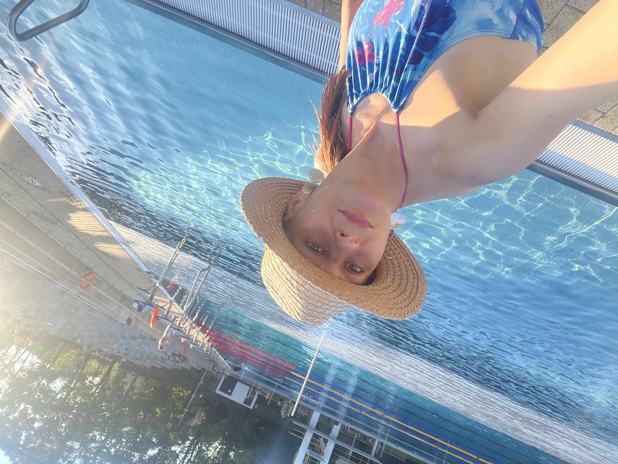 A woman wearing a straw hat and a blue floral swimsuit is taking a selfie by a swimming pool. She is standing in the shallow end of the pool, with the water up to her chest. The pool is large and appears to be an outdoor public pool, with lanes marked and a diving board in the background. There are other people in the background, some swimming and others relaxing by the pool. The sun is shining, and the water is sparkling. The scene appears to be a sunny day at a recreational facility.