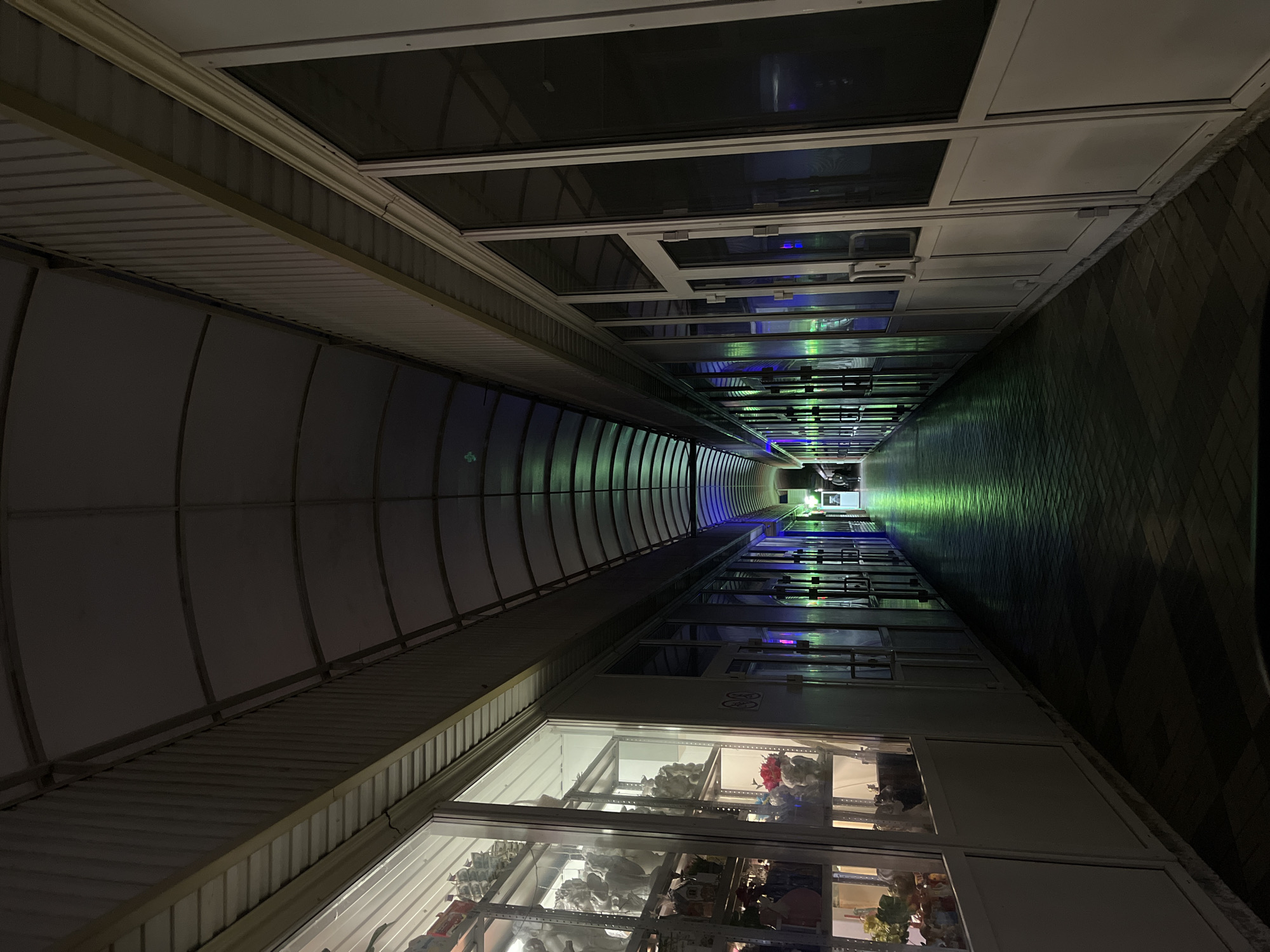 The image depicts a long, narrow, covered passage or corridor, viewed from one end looking towards the distant vanishing point. The scene appears to be indoors or under a protective structure, possibly at night or in low light conditions, given the reliance on artificial lighting.
On the left side, the structure features a curved, corrugated metal wall and ceiling, supported by visible beams. The ceiling above this section appears to have both solid, opaque panels and some transparent or translucent sections, possibly skylights.
The right side of the passage is lined with multiple storefronts or display booths, each enclosed by white-framed glass windows. These windows showcase various items, including what appear to be stuffed animals, figurines, and other decorative goods on shelves within the shops. Reflections of the corridor's lighting are visible on the glass surfaces. The floor on the right side is patterned with dark and lighter square tiles.
Further down the passage, the primary illumination comes from artificial lights, casting distinct blue and green hues that create a glowing effect on the floor and walls, emphasizing the depth and length of the corridor. The colorful lighting reflects off the floor and the windows of the shops.
No people are visible in the image, contributing to a quiet, almost deserted atmosphere. On one of the storefront windows on the right, a small white sign with a black circle and diagonal line, resembling a "no smoking" symbol, is visible. Below it, there is another faint marking that could be a number, possibly "83" or "B3". The overall setting suggests a commercial arcade, covered market, or a pedestrian passage in Krasnodar, Russia.