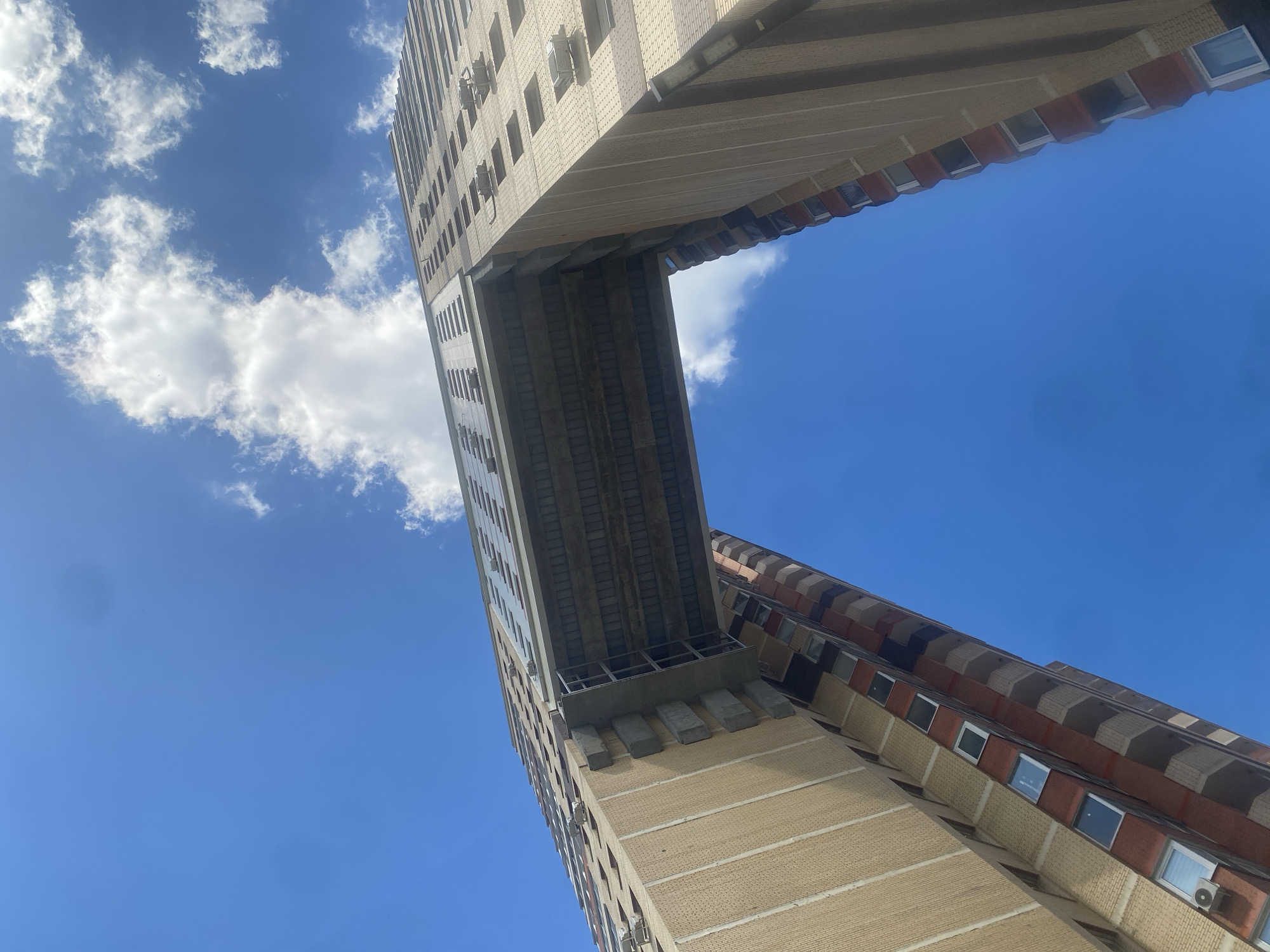 The image shows a low-angle view of a unique architectural structure: two tall residential buildings in Kotelniki, Russia, connected by a skybridge. The buildings are primarily constructed with light-colored brickwork and feature numerous windows. The skybridge appears to be a concrete and metal structure, spanning between the upper floors of the two buildings, creating an open space beneath it. The sky is a bright blue with scattered white clouds. The perspective emphasizes the height and scale of the buildings.
