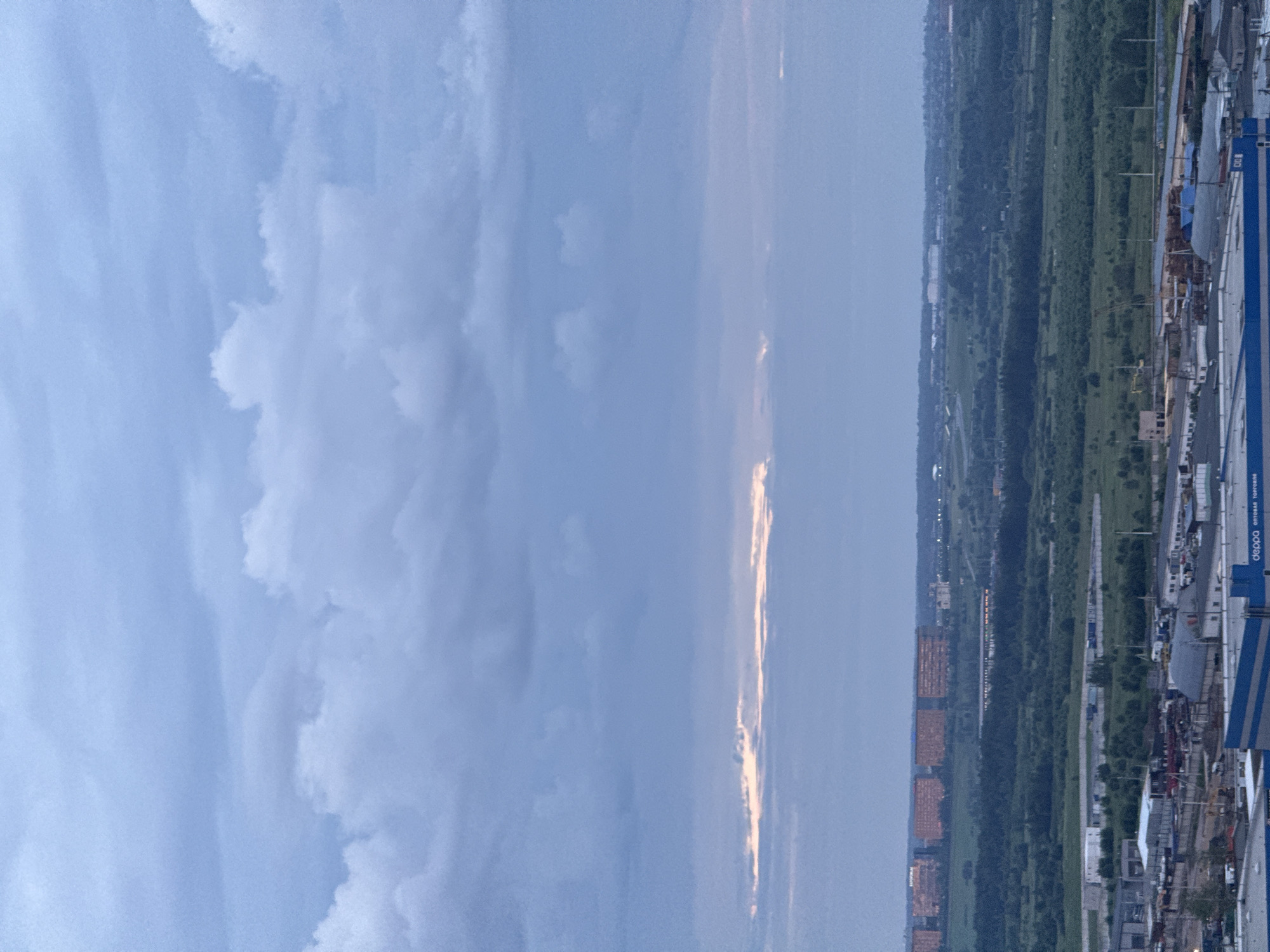 An aerial, wide shot shows a predominantly overcast sky with layered clouds of varying density. A thin, horizontal band of lighter sky, possibly indicating the horizon or a break in the clouds, is visible in the mid-ground, tinged with a warm, orange hue. Below this, a vast, green landscape unfolds, characterized by dense foliage and scattered trees, suggesting a rural or semi-rural area bordering an urban expanse. In the far distance, a row of multi-story buildings, likely residential or commercial, stands out against the darker vegetation. Closer to the foreground, an industrial or commercial zone is discernible, with various structures, including warehouses, smaller buildings, and what appear to be storage areas. The presence of roads and what could be construction materials or vehicles suggests ongoing activity. The overall lighting is dim, consistent with dusk or a heavily clouded day.