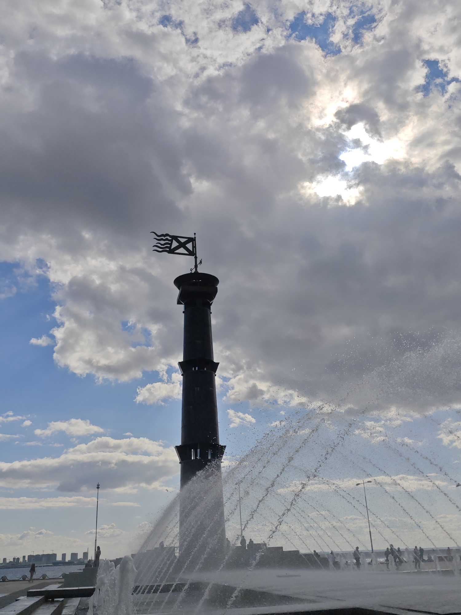 The image depicts a tall, dark, cylindrical tower with a decorative flag at its apex, silhouetted against a dramatic sky filled with both bright sunlight peeking through clouds and large, dark storm clouds. In the foreground, a fountain sprays water in a wide arc, creating a shimmering, curtain-like effect. The tower is located on an embankment or plaza near a body of water, with a cityscape of buildings visible in the distance. Several streetlights and people are scattered along the waterfront promenade. The overall scene suggests a dynamic, potentially windy day in Saint Petersburg, Russia, with the fountain adding a playful element to the grand architecture and the impressive sky.