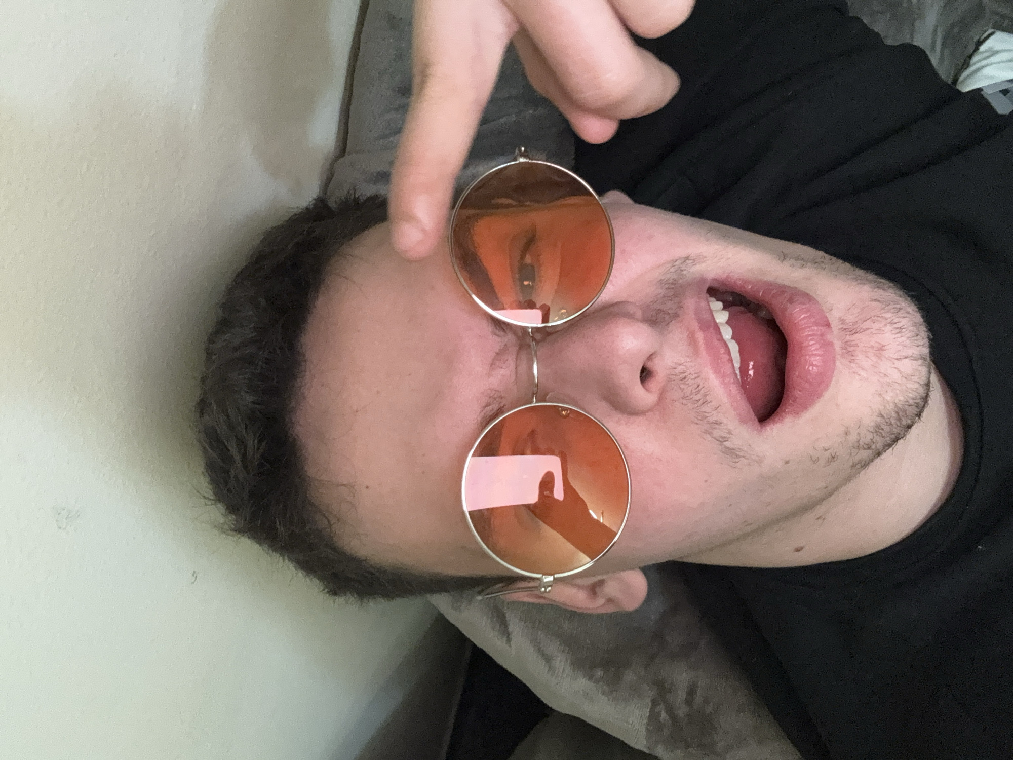 A person is lying down, wearing round, orange-tinted glasses. Their mouth is open. A finger is pointing at the glasses. They are wearing a black shirt. The background includes a wall and a grey textured surface. The location is not discernible from the image.