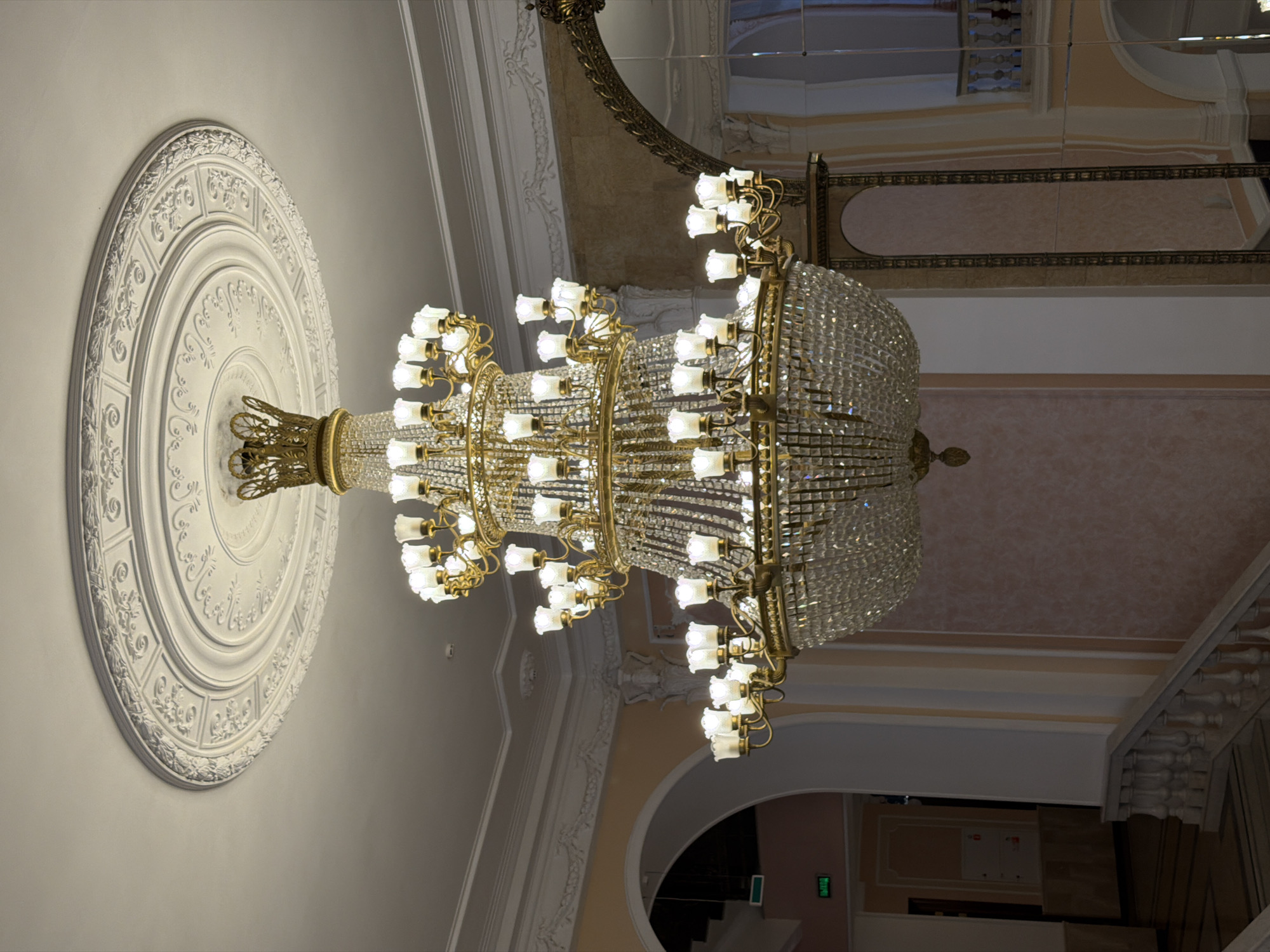 This image captures an ornate, multi-tiered crystal chandelier hanging from a ceiling adorned with decorative plasterwork. The chandelier is predominantly gold and features numerous lights with frosted glass shades, interspersed with crystal elements that reflect light. To the left, a circular, white plaster medallion with intricate floral and geometric patterns is mounted on the ceiling. The background reveals architectural details of what appears to be an interior space, possibly a grand hall or theater, with cream-colored walls and decorative molding. A reflective surface, likely a mirror or polished ceiling, is visible at the top of the image, reflecting parts of the architecture and the chandelier. The lighting suggests an indoor setting, likely during the evening or in a space where artificial light is prominent. There are no people visible in the frame. A faint green illuminated sign with Cyrillic text is partially visible in the lower right portion of the image. The overall impression is one of opulence and classic interior design.