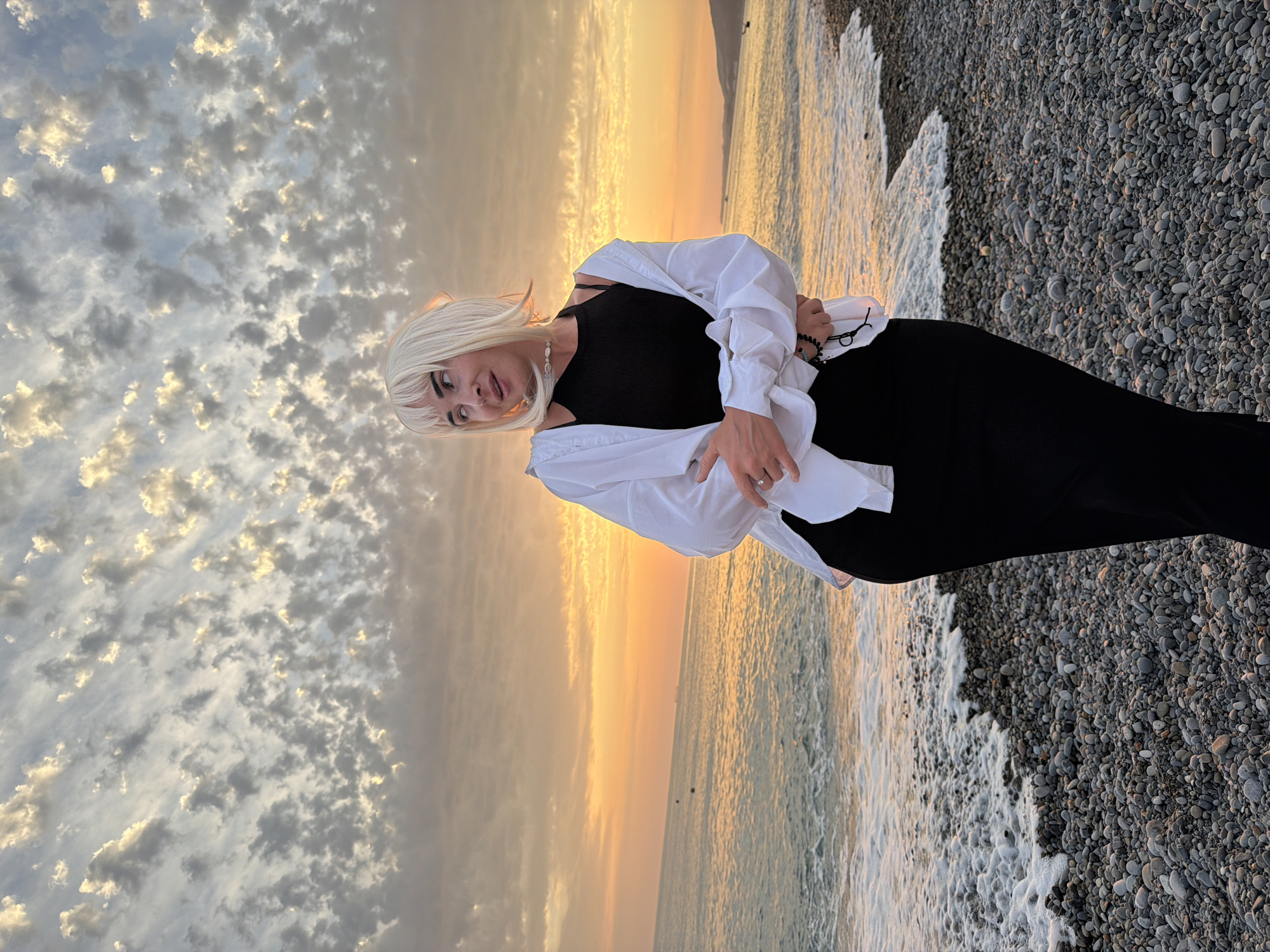 A woman with blonde hair, wearing a black dress and an open white shirt, stands on a pebble beach at sunset. The sky is filled with textured clouds illuminated by the orange glow of the setting sun. The sea gently laps at the shore, with white foam visible on the pebbles. The woman is looking away from the camera, with her arms crossed. The scene appears to be located on a beach in Tuapse, Russia, given the time of day and atmospheric conditions suggesting a coastal location with a pebble shore.