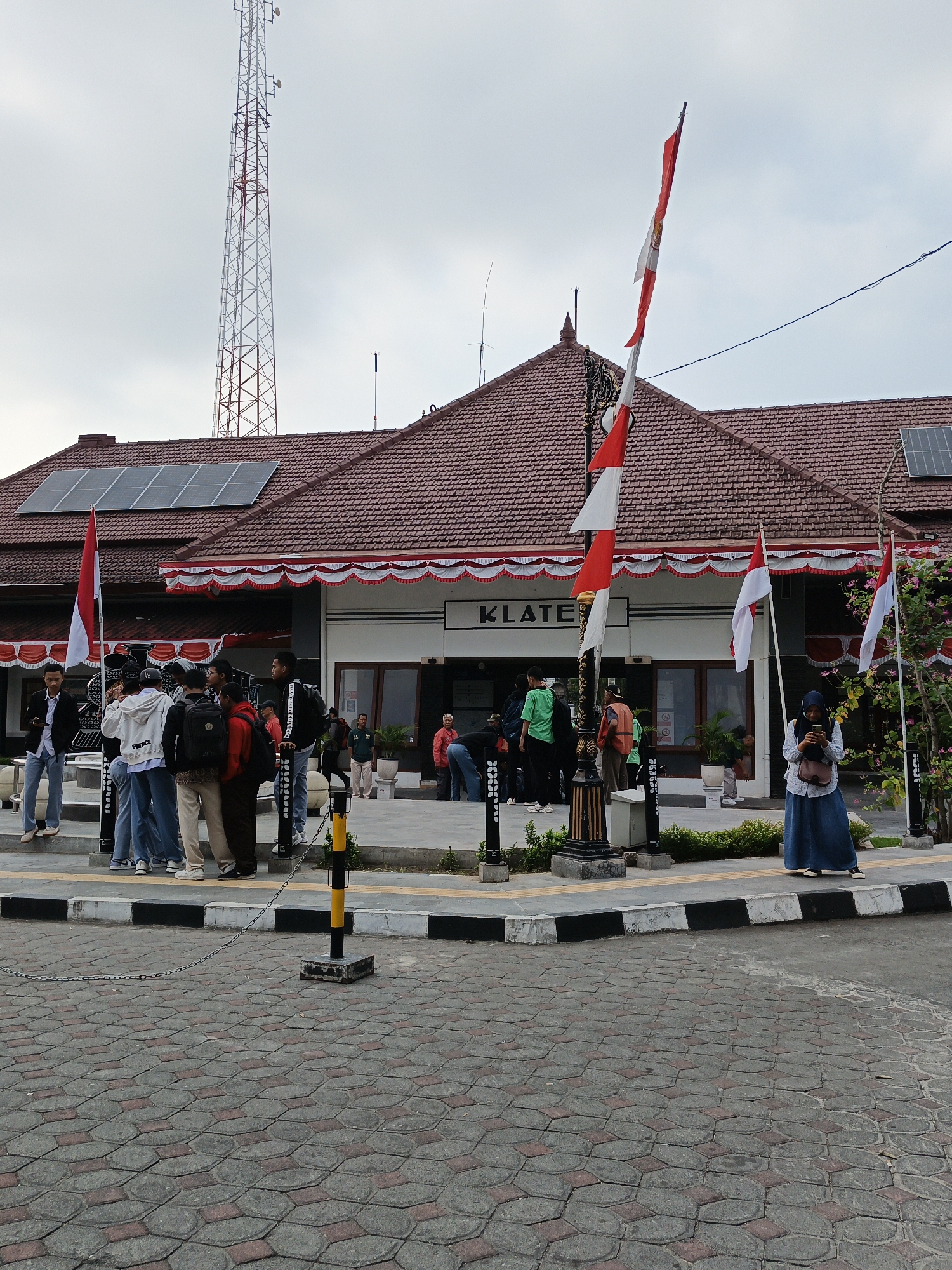The image depicts the exterior of a building in Klaten, Indonesia, likely a public facility given the presence of Indonesian flags and a communication tower in the background. A group of young people, appearing to be students, are gathered on a raised walkway in front of the building. Some are standing and looking around, while others are engaged in conversation. A woman wearing a hijab and a long blue skirt stands to the right, looking at her phone. The building itself has a tiled roof and a banner displaying the word "KLATEN." Solar panels are visible on the roof. The foreground is paved with interlocking hexagonal bricks. The overall atmosphere suggests a casual gathering or transit point.