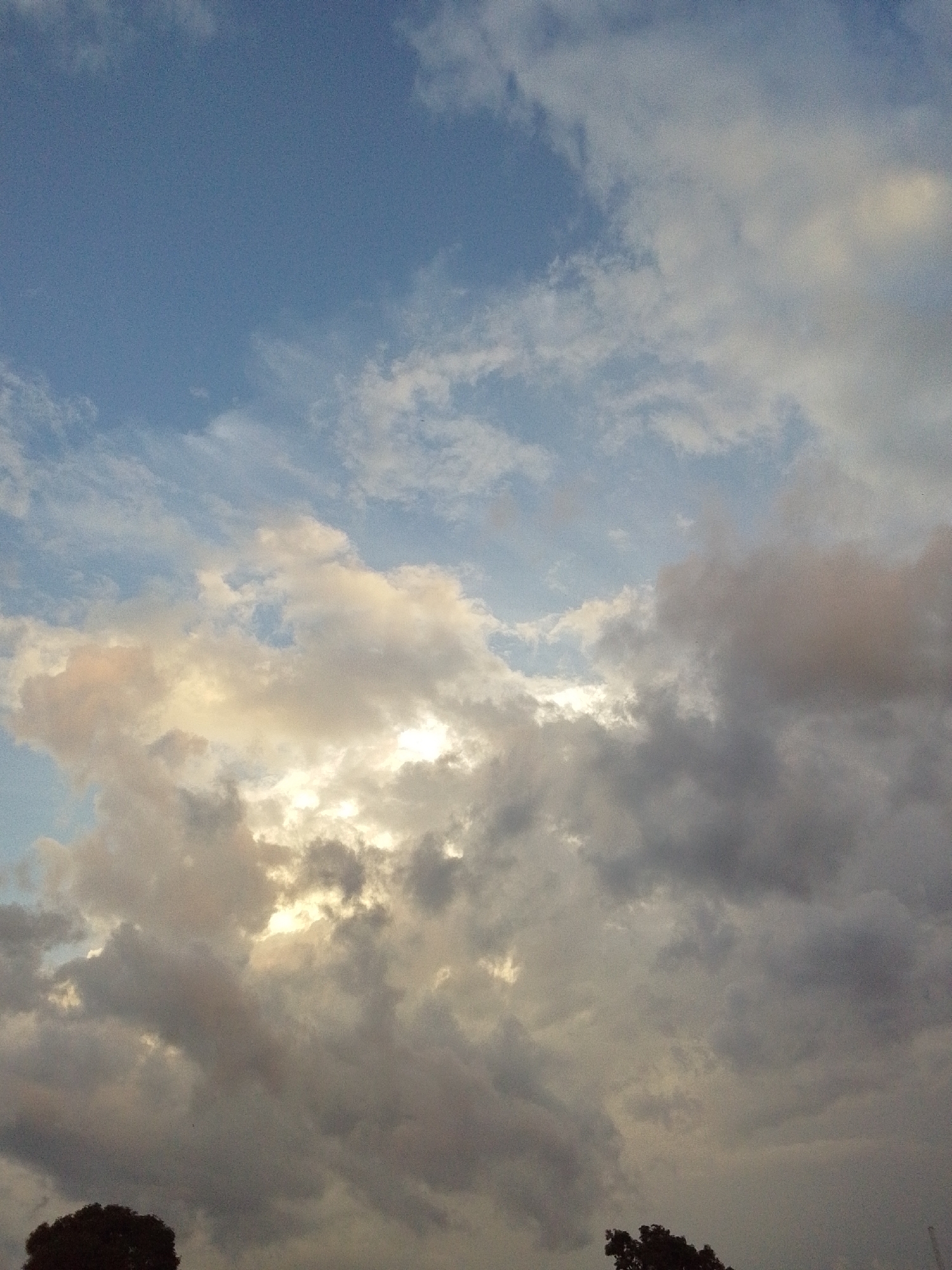 The image depicts a sky with scattered clouds and a sliver of sunlight breaking through. The sky is predominantly blue with patches of white and gray clouds, some appearing dense and others wispy. The sunlight creates a luminous glow in the upper right quadrant of the image, illuminating some of the clouds from behind. At the bottom of the frame, the dark silhouettes of tree branches are visible against the sky. The scene appears to be outdoors, possibly during the late afternoon or early evening. The provided location details indicate the image was taken in Serti, Nigeria.