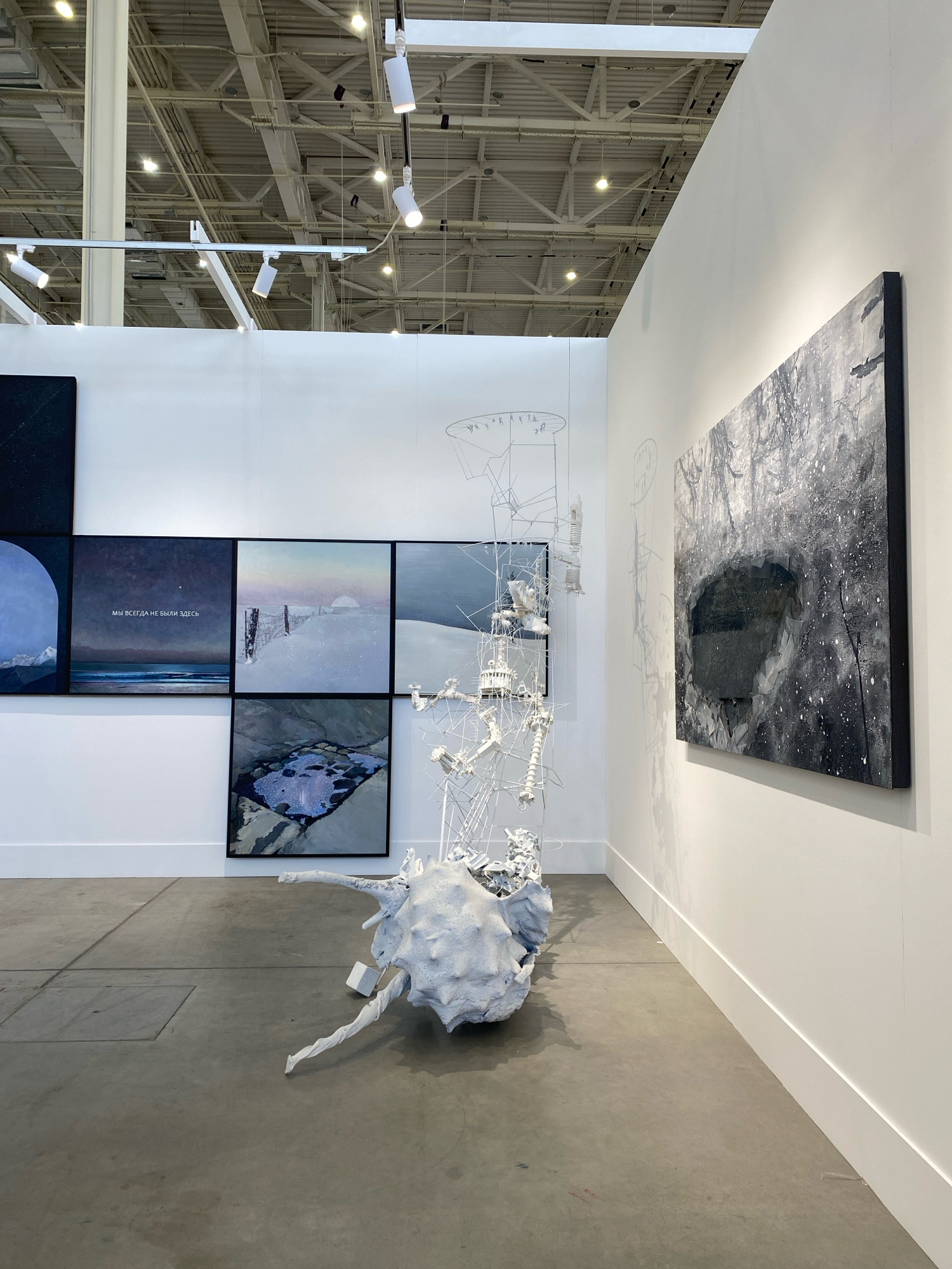 The image depicts an art exhibition space, likely in Khimki, Russia, given the Russian text visible on one of the artworks. The scene features a white-walled exhibition room with a concrete floor. Several pieces of art are displayed. On the left, a grid of square artworks shows abstract and landscape imagery, one of which contains the Russian phrase "МЫ ВСЕГДА НЕ БЫЛИ ЗДЕСЬ" (We were never here). In the center, a large, white, abstract sculpture is positioned on the floor, resembling a skeletal, organic form with spiky protrusions. Suspended from the ceiling above this sculpture is a delicate, wireframe structure. To the right, a large, monochrome framed artwork is mounted on the wall, depicting a dark, textured landscape with a central void. The ceiling above is industrial, with exposed beams and track lighting. The overall impression is of a contemporary art installation within a gallery or exhibition hall.