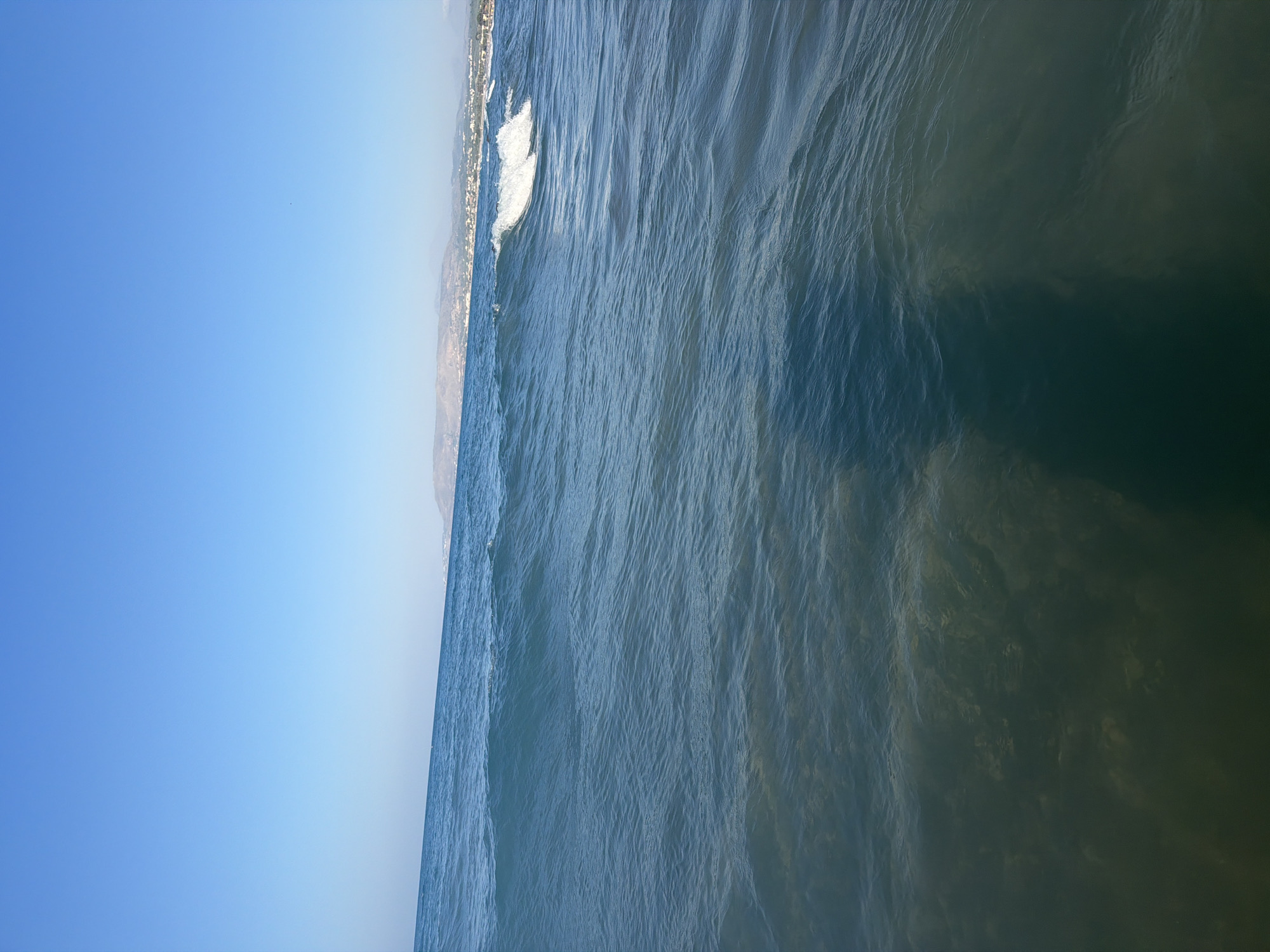 The image depicts a coastal scene in Georgioupoli, Greece. The foreground is dominated by the clear, blue waters of the sea, with gentle waves rolling in. The seabed is visible through the shallow water, appearing sandy and textured. A dark shadow, presumably cast by the photographer, stretches across the lower right portion of the frame. In the midground, a moderate wave is breaking, its white foam contrasting with the deeper blue of the ocean. The horizon line is defined by the meeting of the sea and the sky. In the background, a distant coastline is visible, featuring rolling hills and scattered buildings that suggest a town or resort area. The sky above is a clear, bright blue, indicating a sunny day.