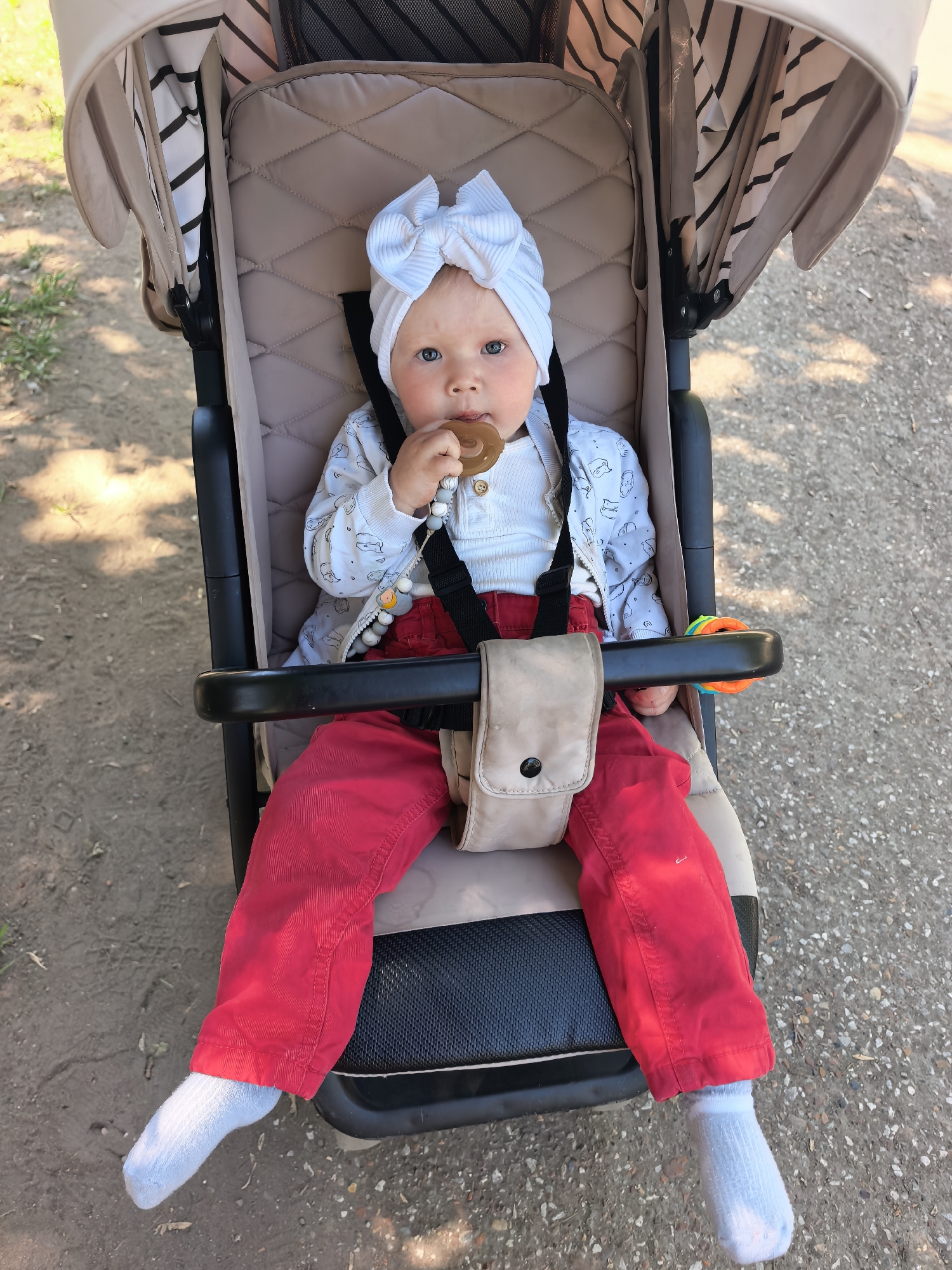 A fair-skinned baby is seated in a stroller, wearing a white ribbed headband with a bow, a white long-sleeved onesie with a patterned jacket, red pants, and white socks. The baby holds a brown pacifier in their mouth. A beaded pacifier clip is visible. The stroller has a beige interior and a canopy with black and white stripes. A beige pouch is attached to the stroller's safety harness. A multi-colored toy is fixed to the stroller's bar. The location appears to be outdoors, potentially in the city of Sterlitamak, Russia, though specific geographic details are not discernible from the image. The ground is a mix of dirt and gravel.