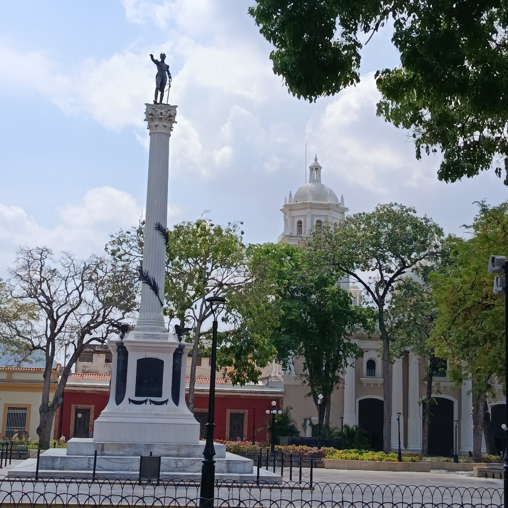 The image shows a statue of a man on a column in a city square. The statue is atop a tall, white column with ornate detailing, and the statue is dark in color, possibly bronze. The man is depicted holding a sword and raising his other arm in what seems like a commanding gesture.
Behind the column and statue, there's a large building with a white dome, partially obscured by trees. The building appears to be a church or a similar type of religious structure, given its architectural style.
In the foreground, there's a black metal fence enclosing the area around the statue. The setting is an urban environment, as suggested by the surrounding buildings and street furniture like lampposts.
The location is the Plaza Bolivar of La Guaira in Venezuela.