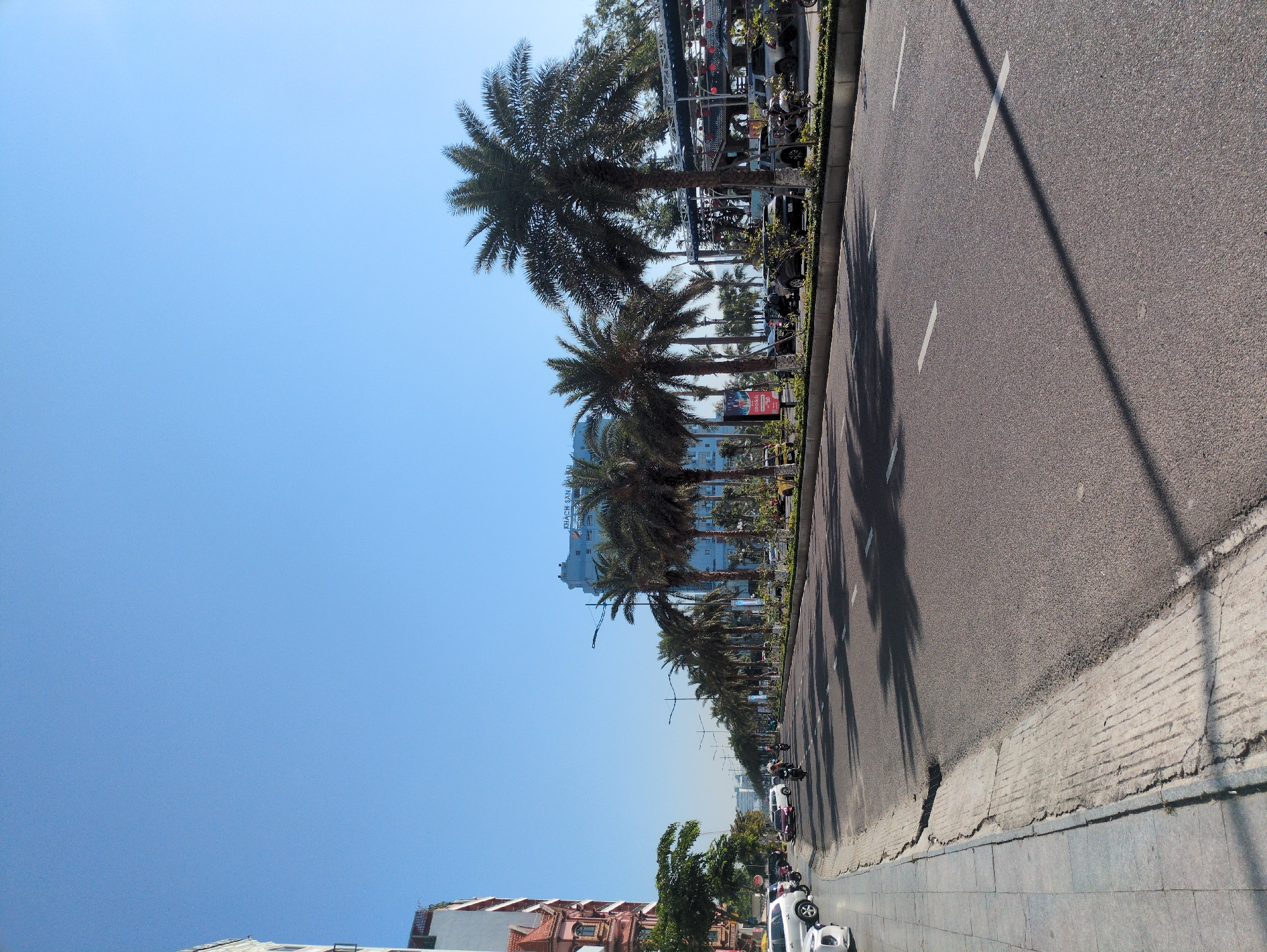 The image depicts a wide, multi-lane urban thoroughfare in South Quy Nhon Ward, Vietnam, under a clear blue sky during daylight hours. The perspective is street-level, looking down the road.
On the immediate left, a concrete sidewalk with visible cracks parallels the road. Several vehicles are parked along this curb, including multiple white and silver sedans and SUVs, and a red scooter. Further left, a multi-story building with reddish-brown and white architectural elements is partially visible, alongside green foliage.
The road itself is paved with dark asphalt, featuring clear white dashed lane markings. Sparse traffic is present, with multiple motorcycles observed moving away from the camera, one ridden by an individual. Distinct, elongated shadows from palm trees and streetlights extend across the road from the right.
Separating the roadway is a landscaped median densely lined with mature palm trees, their dark green fronds casting significant shadows. Green shrubs are planted at the base of these trees, and tall, dark streetlights are interspersed along the median.
In the mid-ground and background on the right, a prominent blue multi-story building, identified by the text "KHÁCH SẠN SAN HÔ" (Coral Hotel) on its roofline, stands behind the palm trees. Adjacent to this and further down the road, a semi-covered commercial or dining area is visible, featuring a structure adorned with red lanterns and string lights, surrounded by numerous parked cars and motorcycles. Distant buildings are faintly discernible on the horizon.