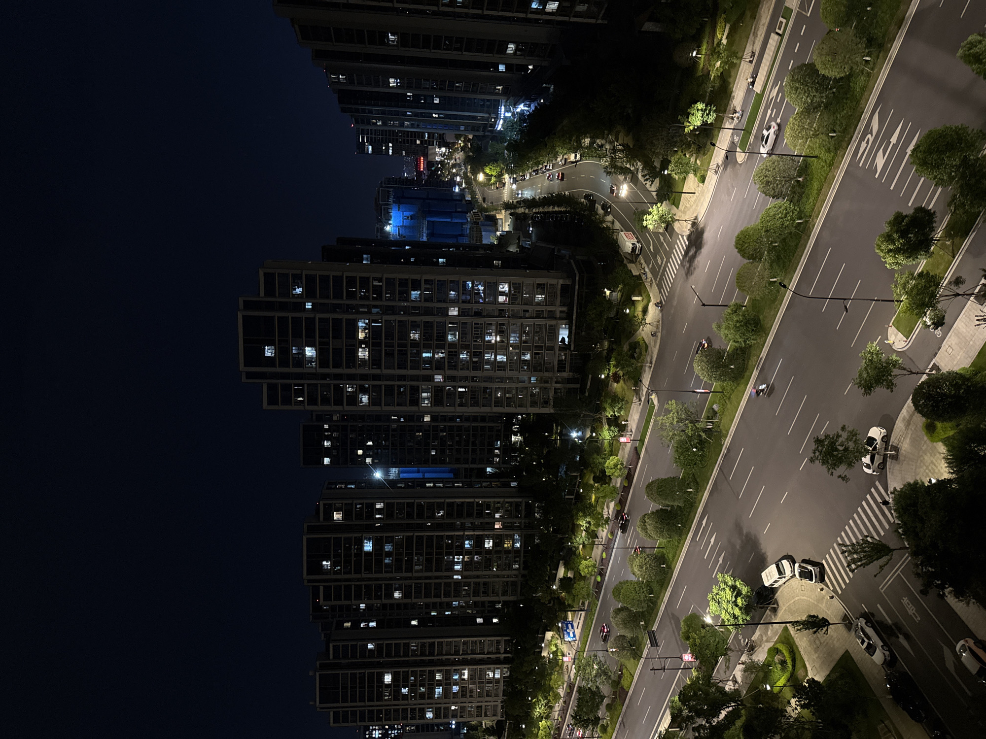 This is a nighttime aerial view of a modern urban intersection in Yiwu, China. The scene is dominated by several tall residential apartment buildings with many lit windows, suggesting occupancy. A multi-lane road runs through the center of the frame, with streetlights illuminating the asphalt. Several vehicles, including cars and what appears to be a delivery truck, are visible on the road, some in motion and others parked. Pedestrian crosswalks are marked on the road surface. Lush green trees and landscaping line the sides of the road and the bases of the apartment buildings. The sky is dark, indicating it is nighttime. The architecture and the presence of high-rise buildings are characteristic of a developed urban area.