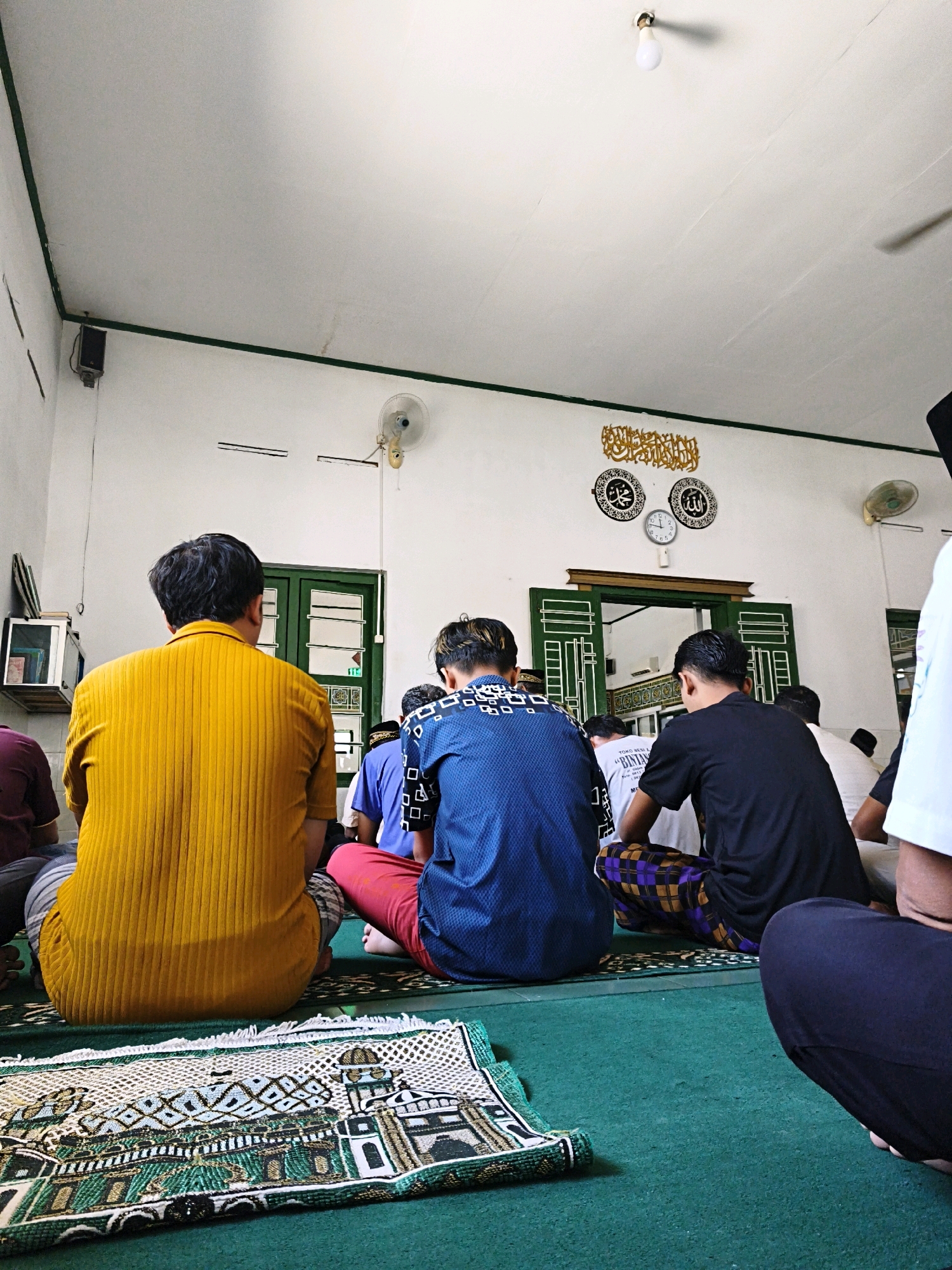 This image depicts an indoor scene within a mosque in Desa Dibal, Indonesia, where several men are engaged in prayer. The perspective is from behind the individuals, focusing on their backs as they sit on a green carpeted floor, facing towards the front of the room. In the foreground, a prayer mat with an intricate design featuring a mosque is laid out. The men are dressed in various casual attire, including a bright yellow long-sleeved shirt, a blue patterned shirt, and a black t-shirt, with some wearing sarongs. The walls of the mosque are painted white with green trim along the top. Decorative Islamic calligraphy and two circular ornaments with Arabic script are mounted on the wall above a green-doored entryway. Two ceiling fans are visible, one near the corner and another on the wall. A light fixture with a single bulb hangs from the ceiling. The overall atmosphere suggests a moment of quiet devotion during a prayer service.