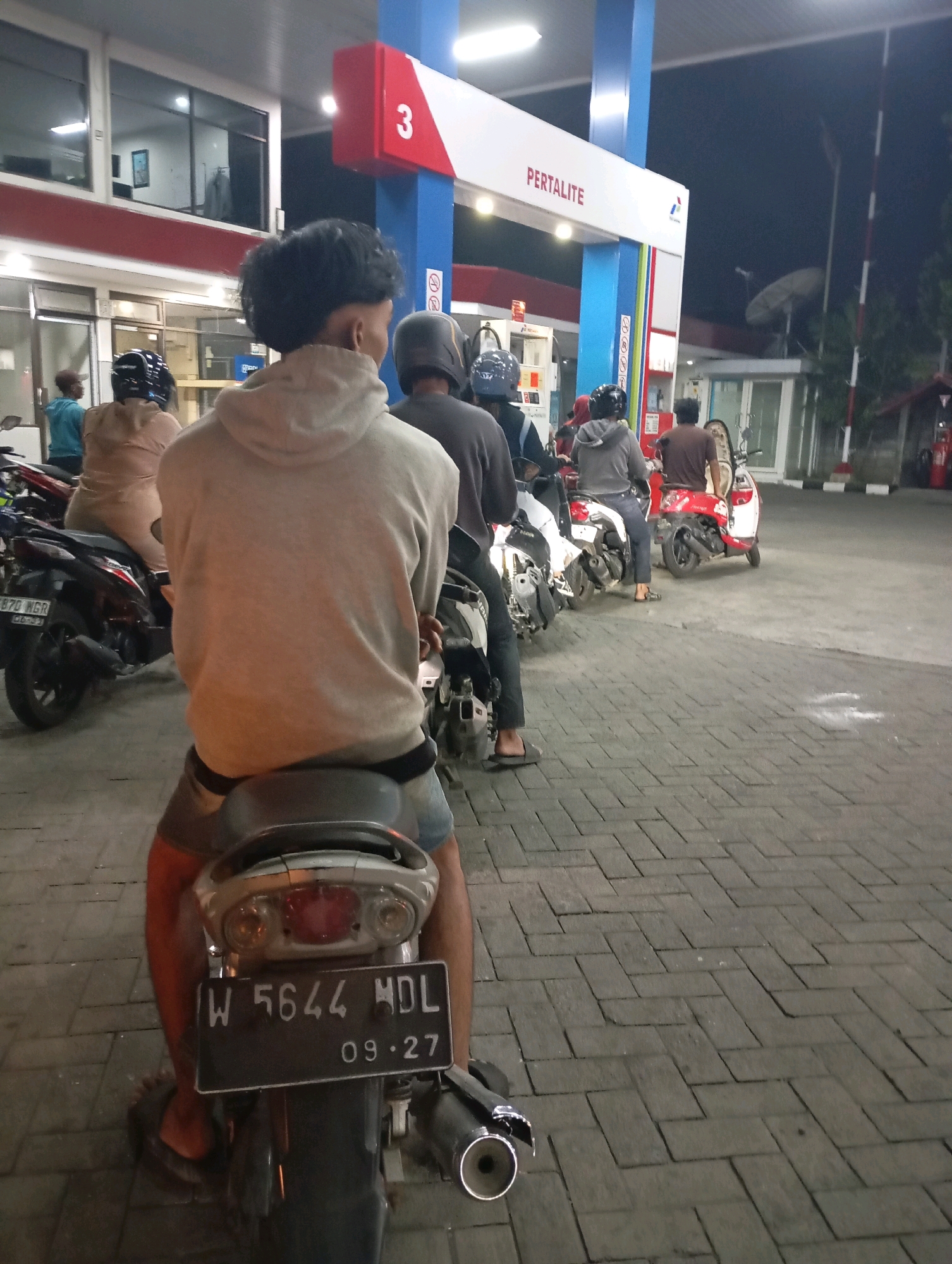 A nighttime scene at a Pertamina gas station in Ketegan, Indonesia, features a queue of motorcycles waiting to refuel. The foreground is dominated by the back of a person on a motorcycle, facing away from the camera, wearing a light-colored hoodie and shorts. The motorcycle's license plate is visible, reading "W 5644 MDL 09-27". Other individuals on motorcycles are visible further back in the queue. The gas station canopy, illuminated by lights, displays the "PERTAMALITE" brand. The ground is paved with interlocking brick.
