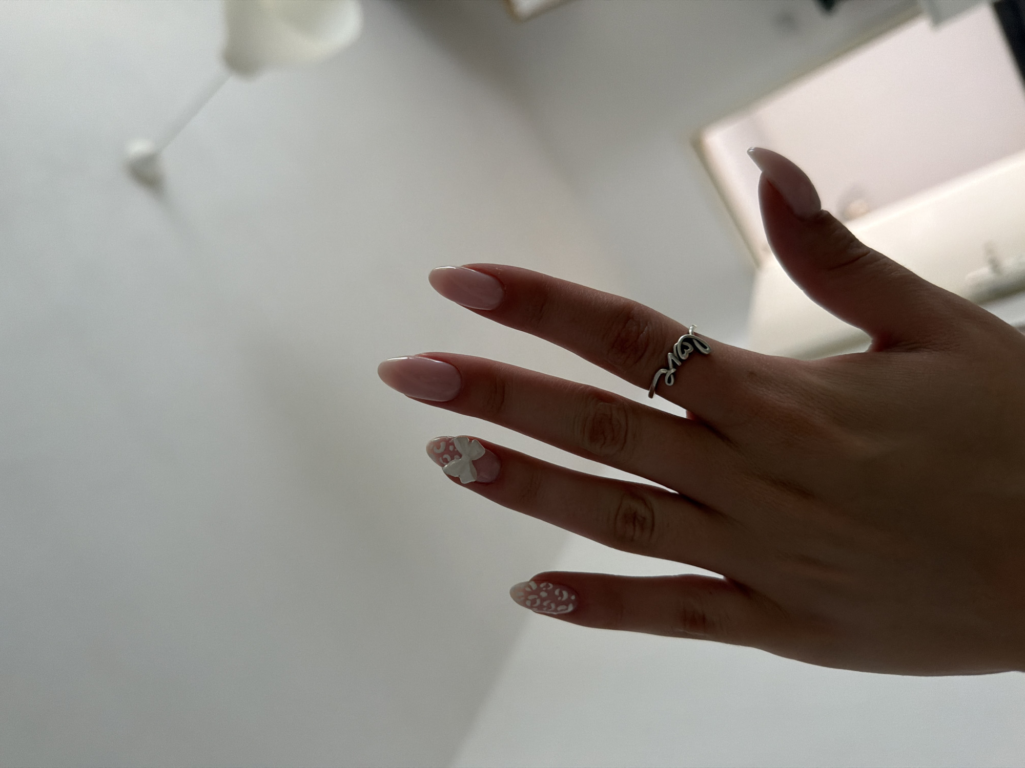 The image displays a close-up view of a person's left hand, with fingers extended upwards, set against a predominantly plain, light-colored background. The hand exhibits a fair skin tone. All visible fingernails are long, with an oval or almond shape, and feature a light pinkish-nude base coat. The nail on the middle finger is adorned with a prominent white three-dimensional bow design. The nail on the index finger displays a pattern of multiple small, abstract white shapes, resembling leopard spots. A silver-toned ring, intricately shaped to spell the word "love" in a cursive font, is worn on the ring finger. The background consists primarily of an undifferentiated white surface, indicative of an indoor ceiling or wall. A blurred white fixture, possibly a light or smoke detector, is faintly visible on the ceiling in the upper right portion of the frame. Towards the bottom right, a faint, blurred outline suggests a doorway or opening with a light-colored frame. The perspective is from below, looking upwards towards the hand and the ceiling. The scene is located in the city of Plovdiv, Bulgaria.